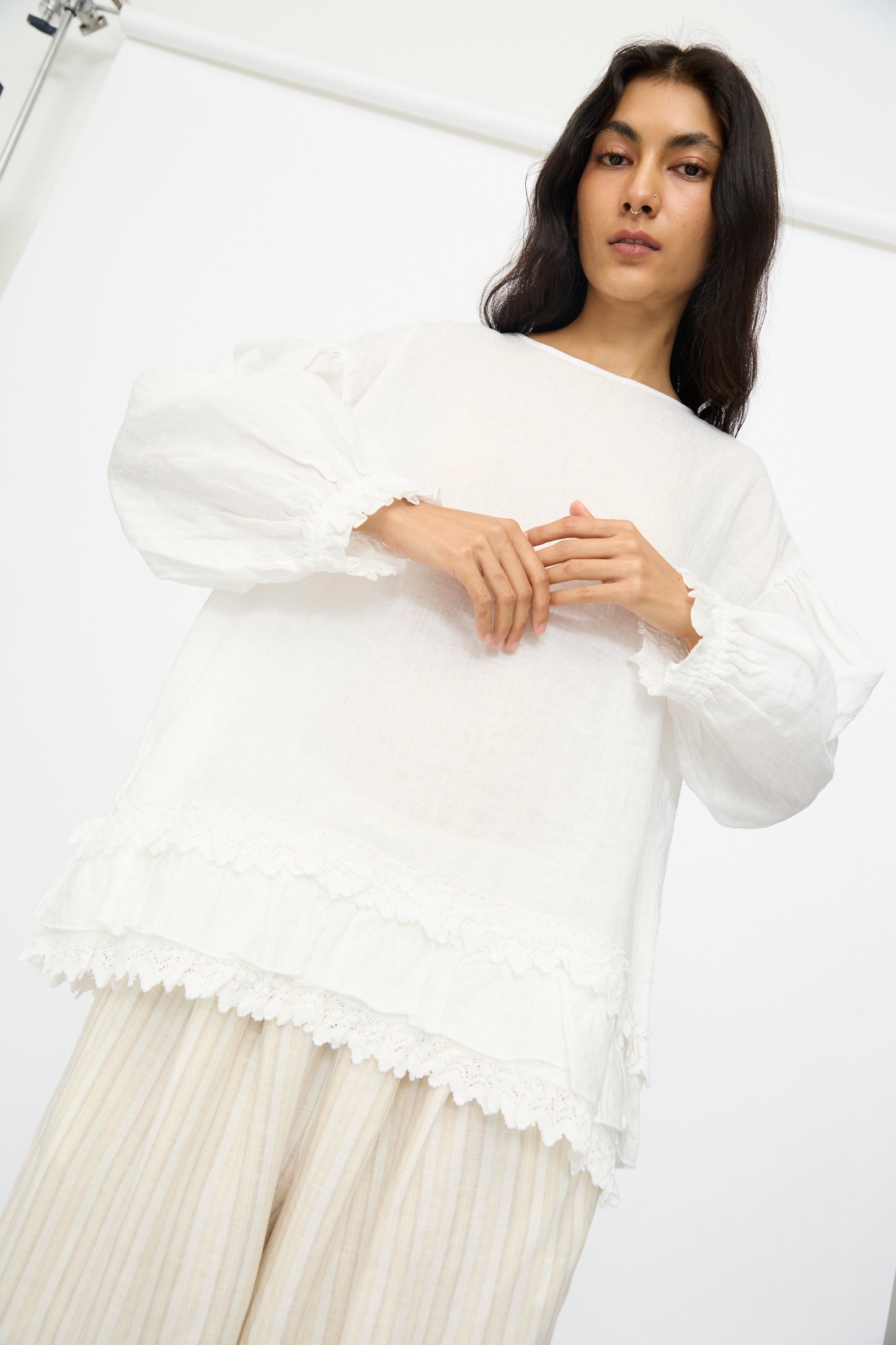 A person in nest Robe's Linen Cotton Lace-Hem Two Way Blouse in Off White and light pleated pants stands against a plain background.