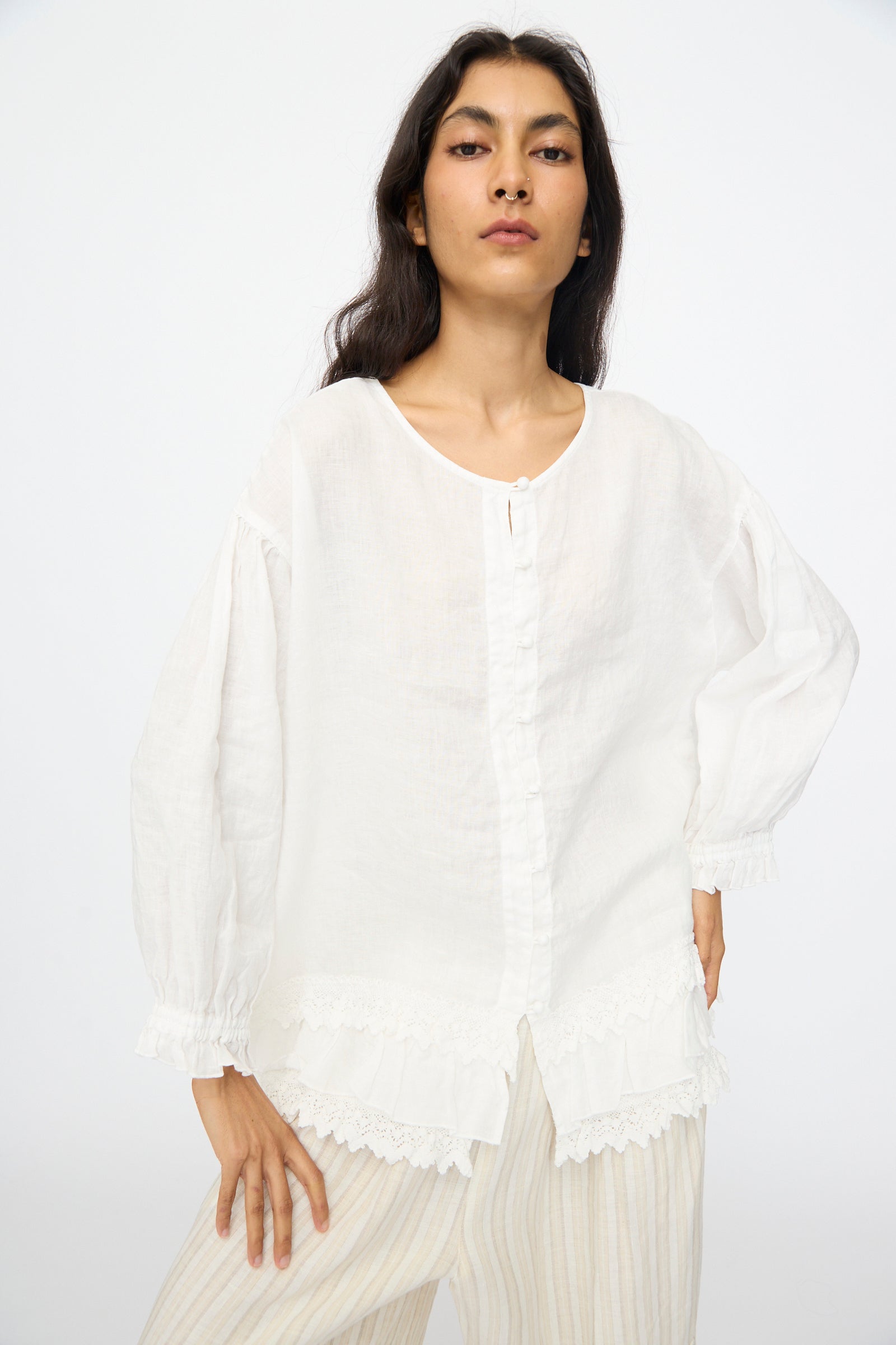 A person wearing the nest Robe Linen Cotton Lace-Hem Two Way Blouse in Off White, featuring cotton lace at the hem and cuffs, and light-colored striped pants, stands against a plain background.