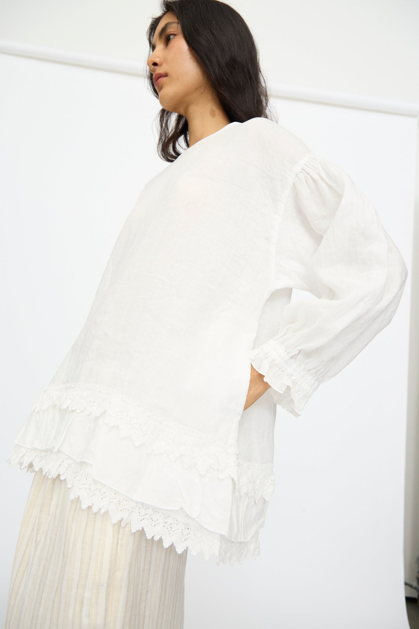 A person with long dark hair wears the nest Robe Linen Cotton Lace-Hem Two Way Blouse in Off White, styled loosely and paired with light pleated pants, standing against a plain white background.