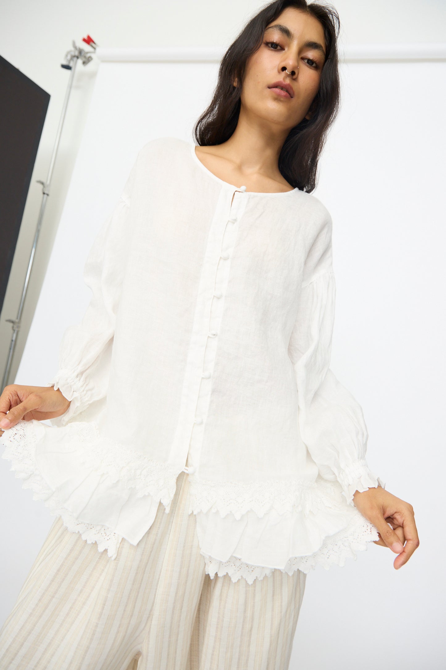 A woman wears the nest Robe Linen Cotton Lace-Hem Two Way Blouse in Off White, styled with light striped pants, standing against a plain white background.
