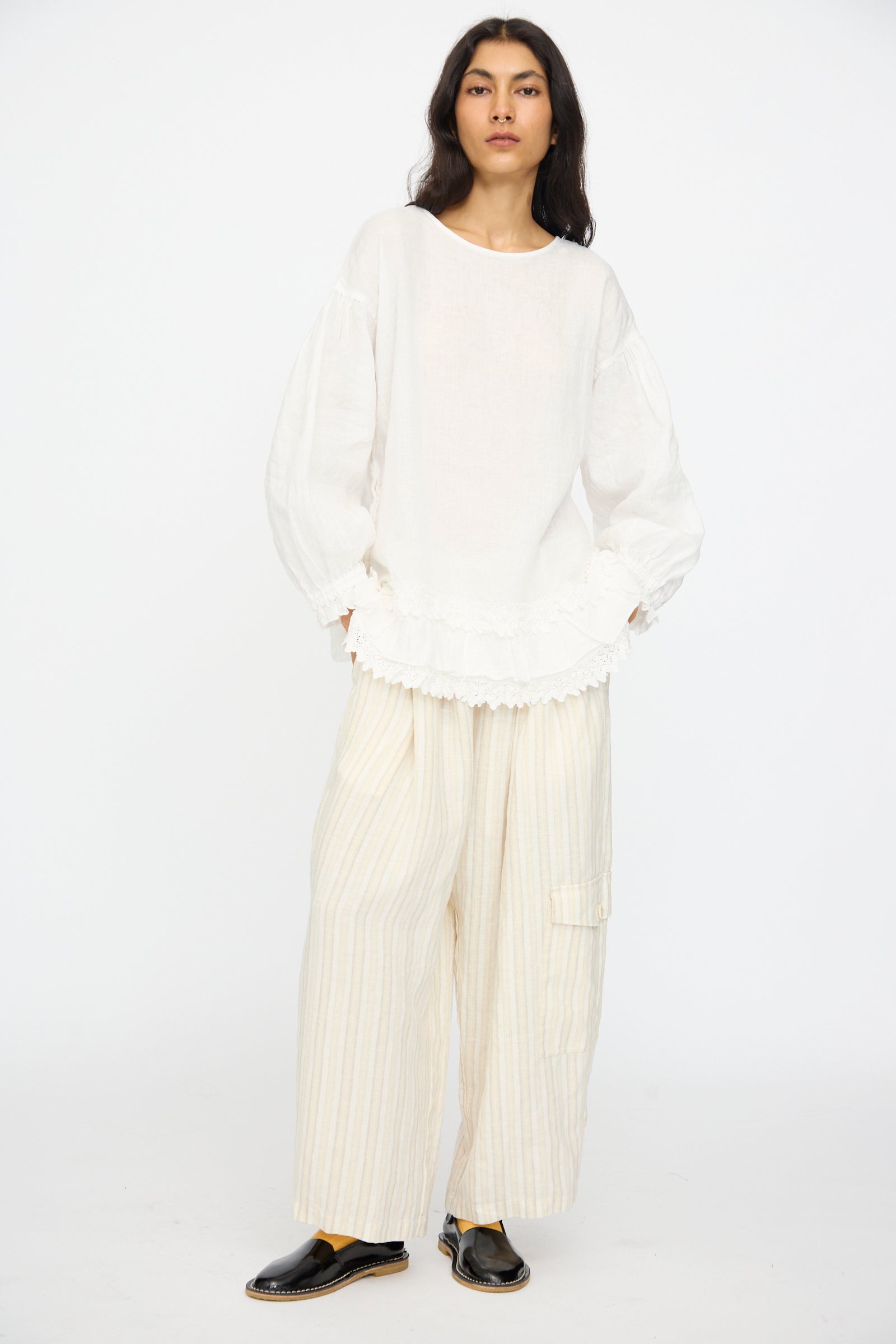 A person stands against a plain background wearing the nest Robe Linen Cotton Lace-Hem Two Way Blouse in Off White, wide cream striped pants, and black slip-on shoes.