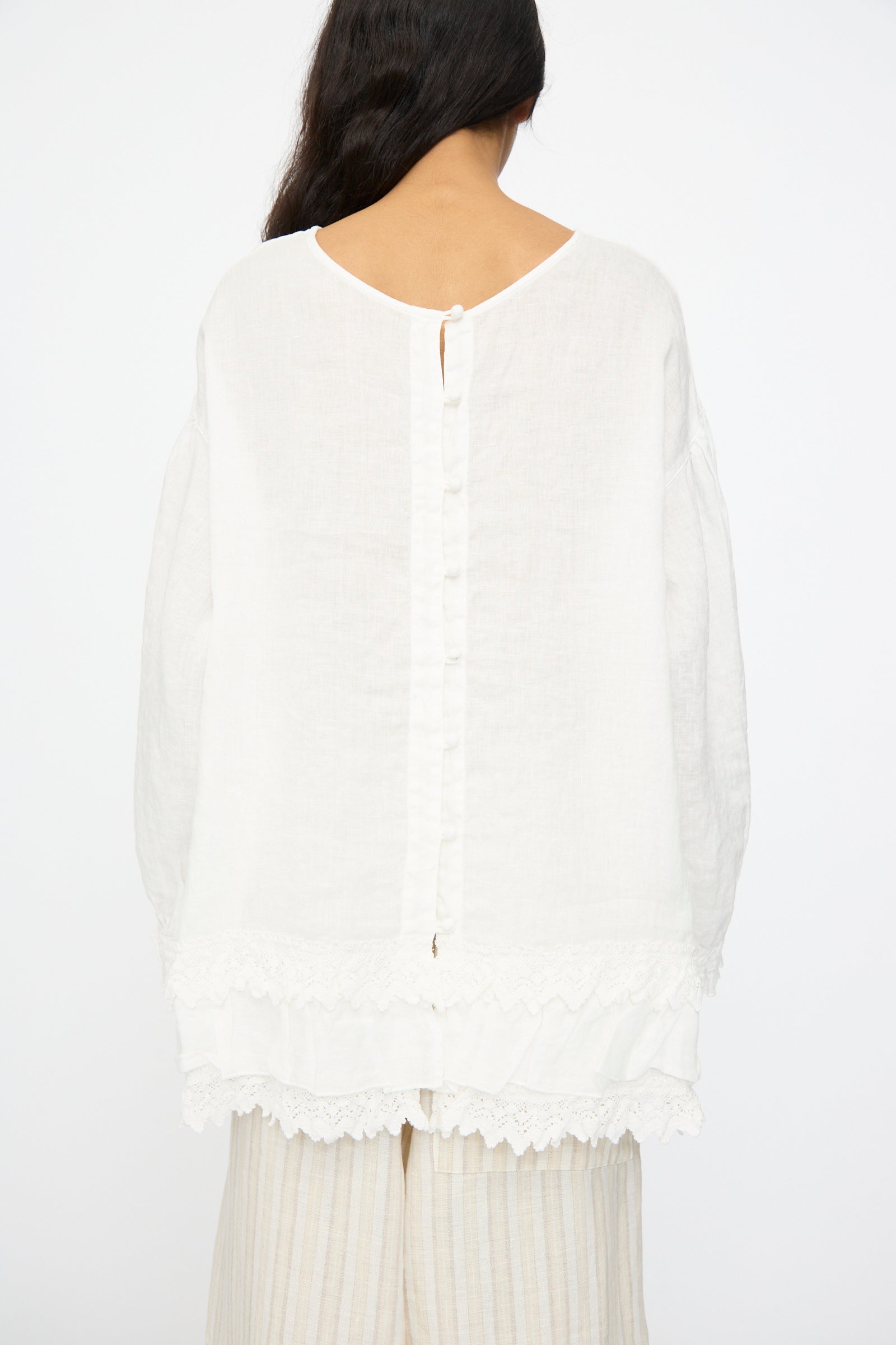 A person wears the nest Robe Linen Cotton Lace-Hem Two Way Blouse in Off White, featuring cotton lace trim and back button details, paired with light striped pants and shown from the back.