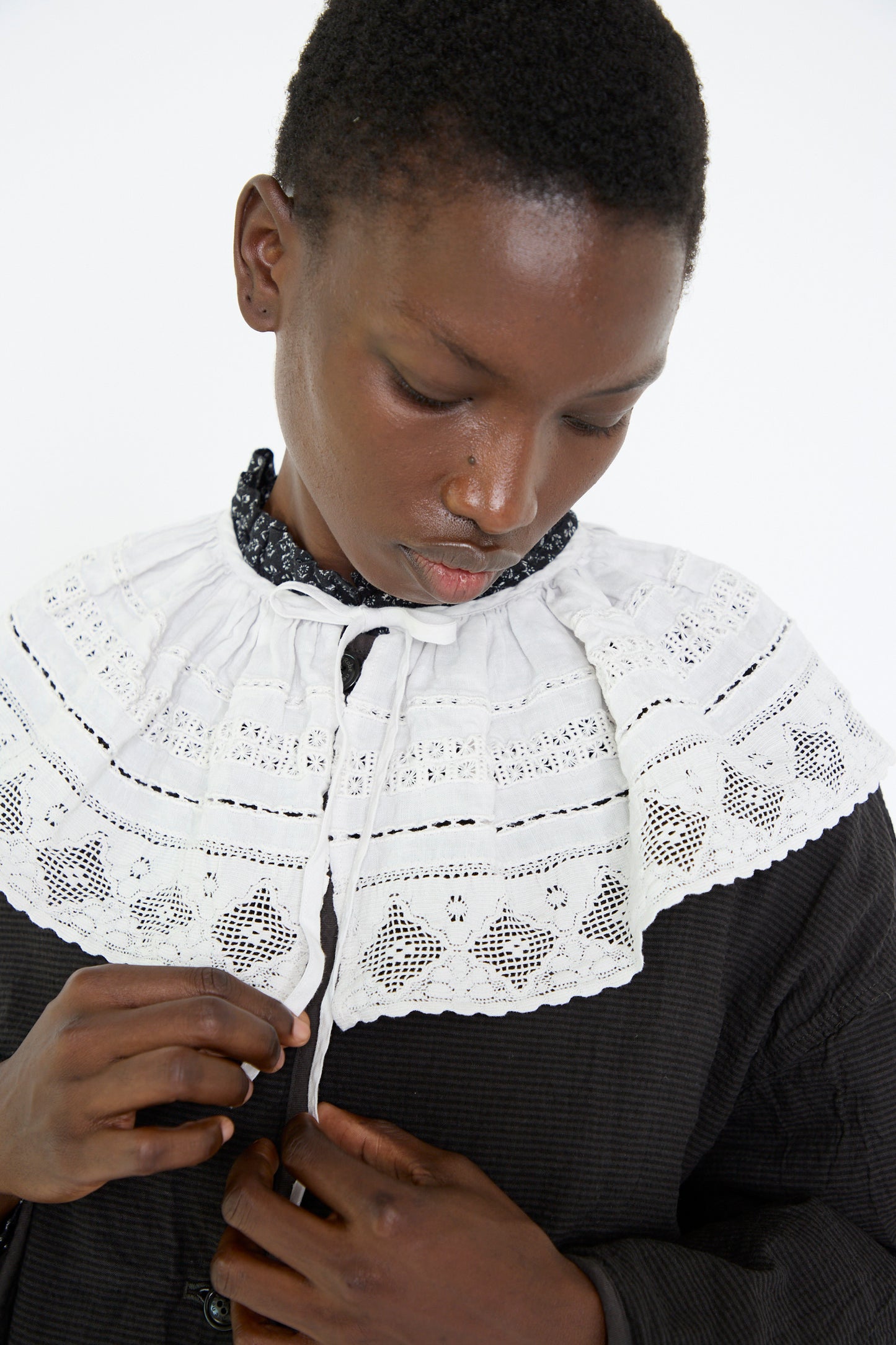 A woman wears the Linen Cotton Lace Detachable Collar in Off White from nest Robe, over a black dress and gazes downward against a plain background.