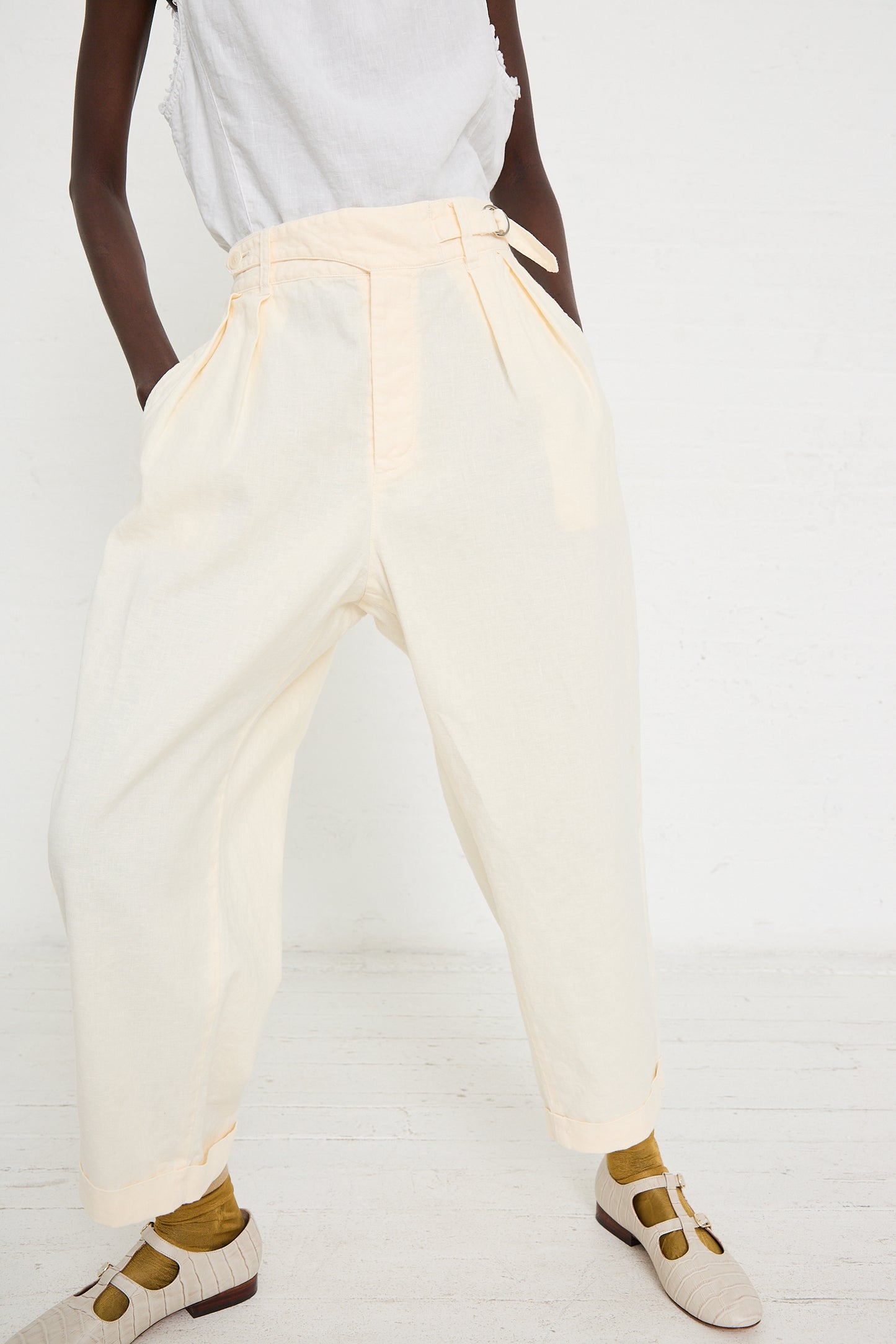 Person wearing light-colored, high-waisted Linen Cotton Blend Gurkah Pant in Off White by nest Robe with pleats and adjustable side tabs, a white top, yellow socks, and beige sandals. Standing against a white background.