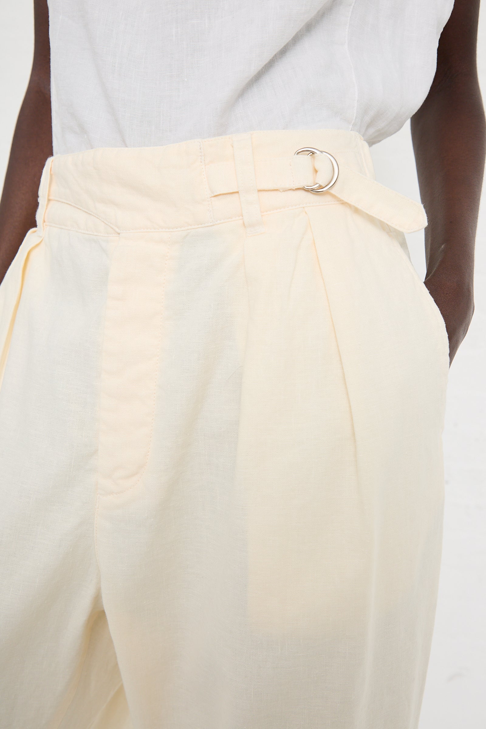 Close-up of a person wearing nest Robe Linen Cotton Blend Gurkah Pant in Off White with a belt and D-ring buckle. Their hands are in the pants' pockets, and they are also wearing a white top.