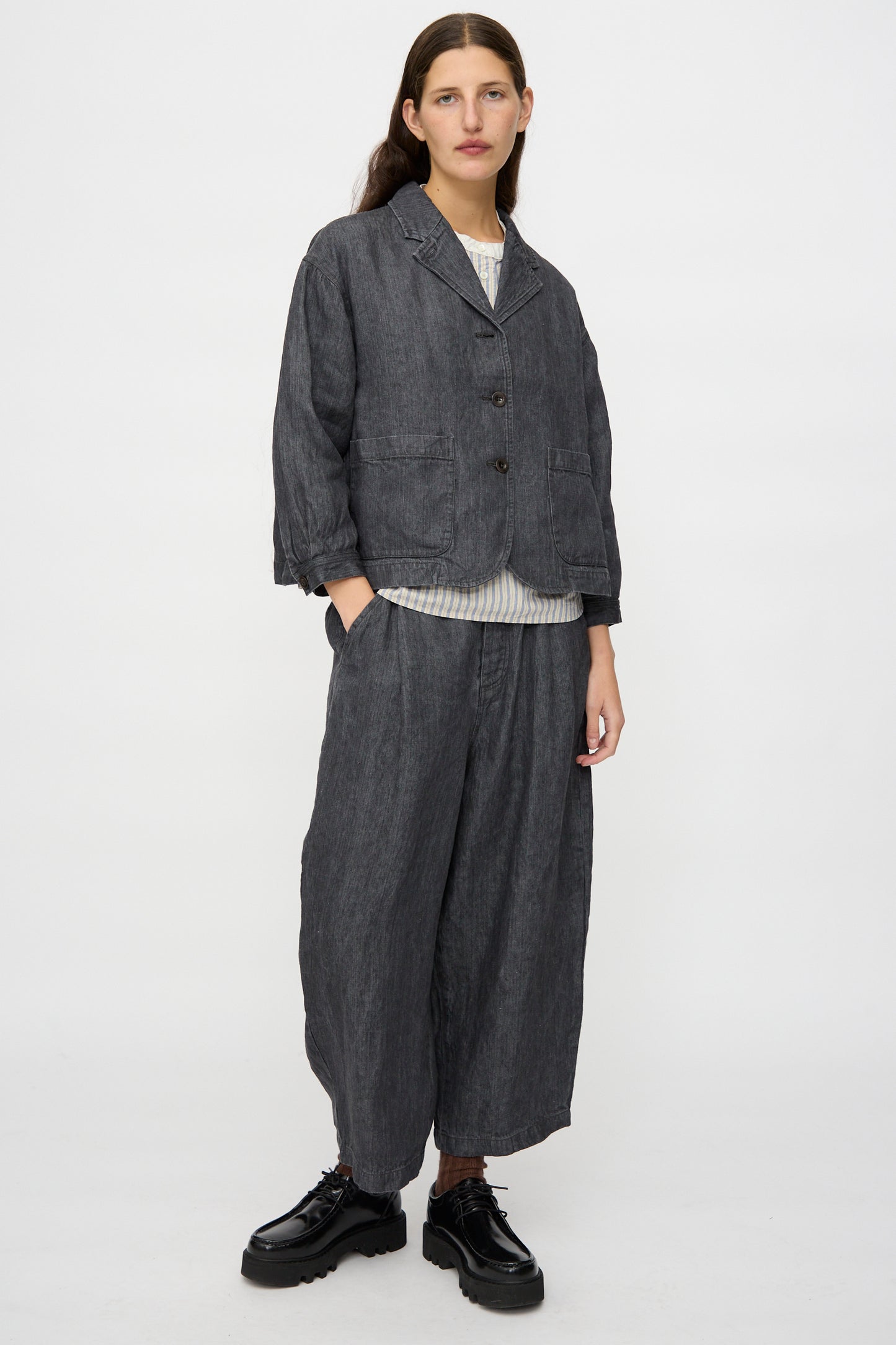 A person stands against a plain background wearing the nest Robe Linen Denim Cropped Jacket in Black, styled with a striped shirt and black platform shoes.