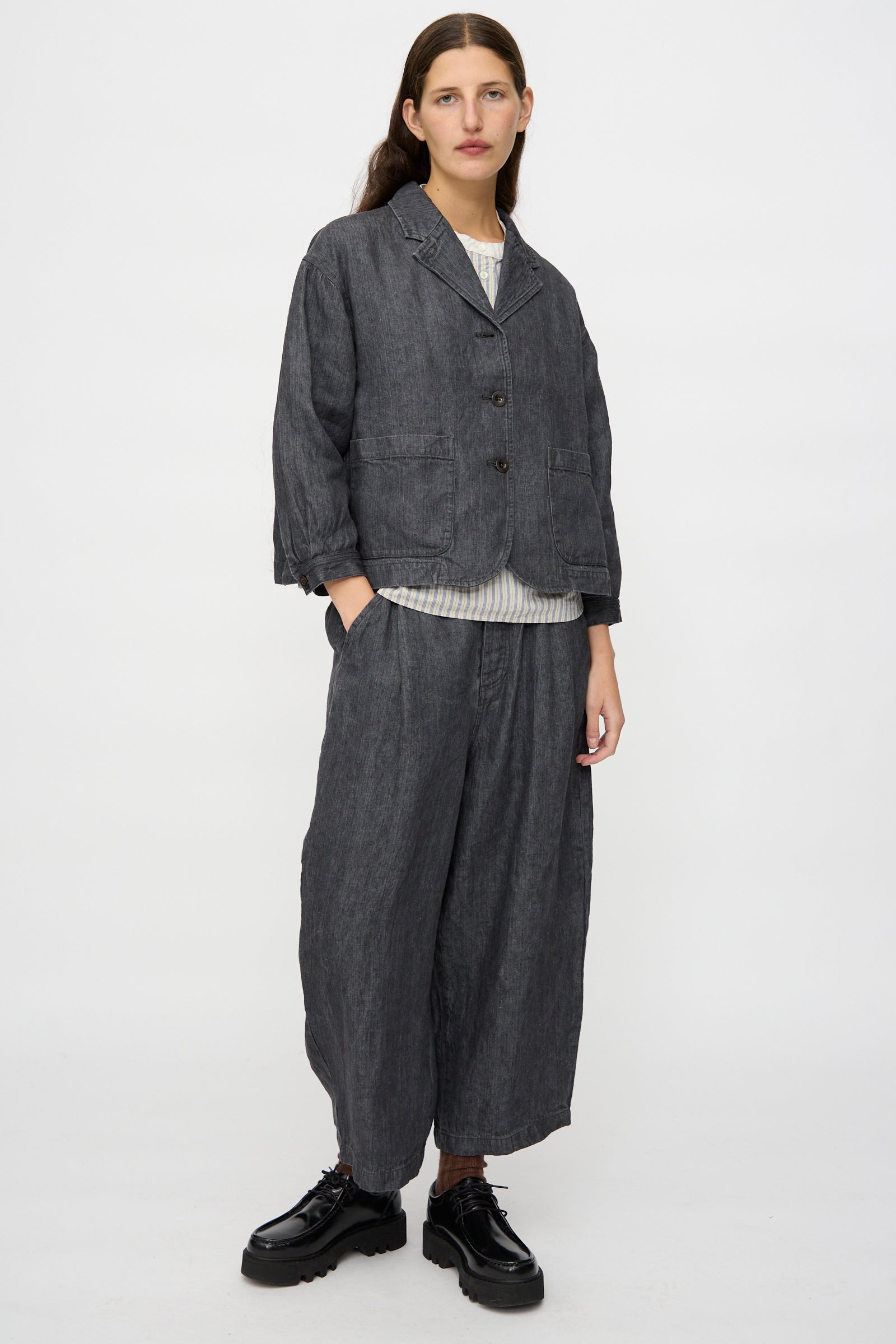 A person stands against a plain background wearing the nest Robe Linen Denim Cropped Jacket in Black, styled with a striped shirt and black platform shoes.