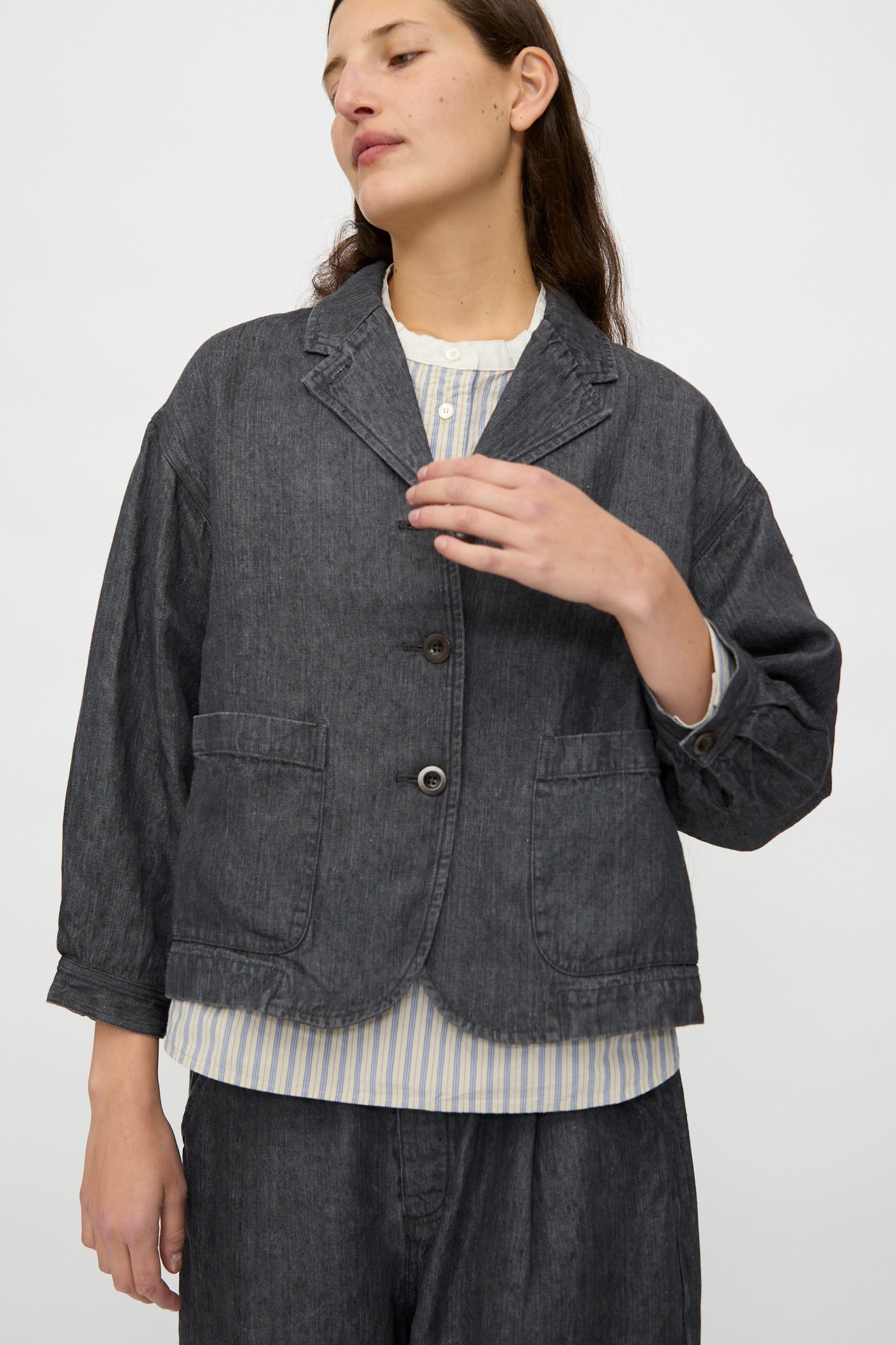 Model wears the nest Robe Linen Denim Cropped Jacket in Black, styled over a light striped shirt, standing against a plain white background.