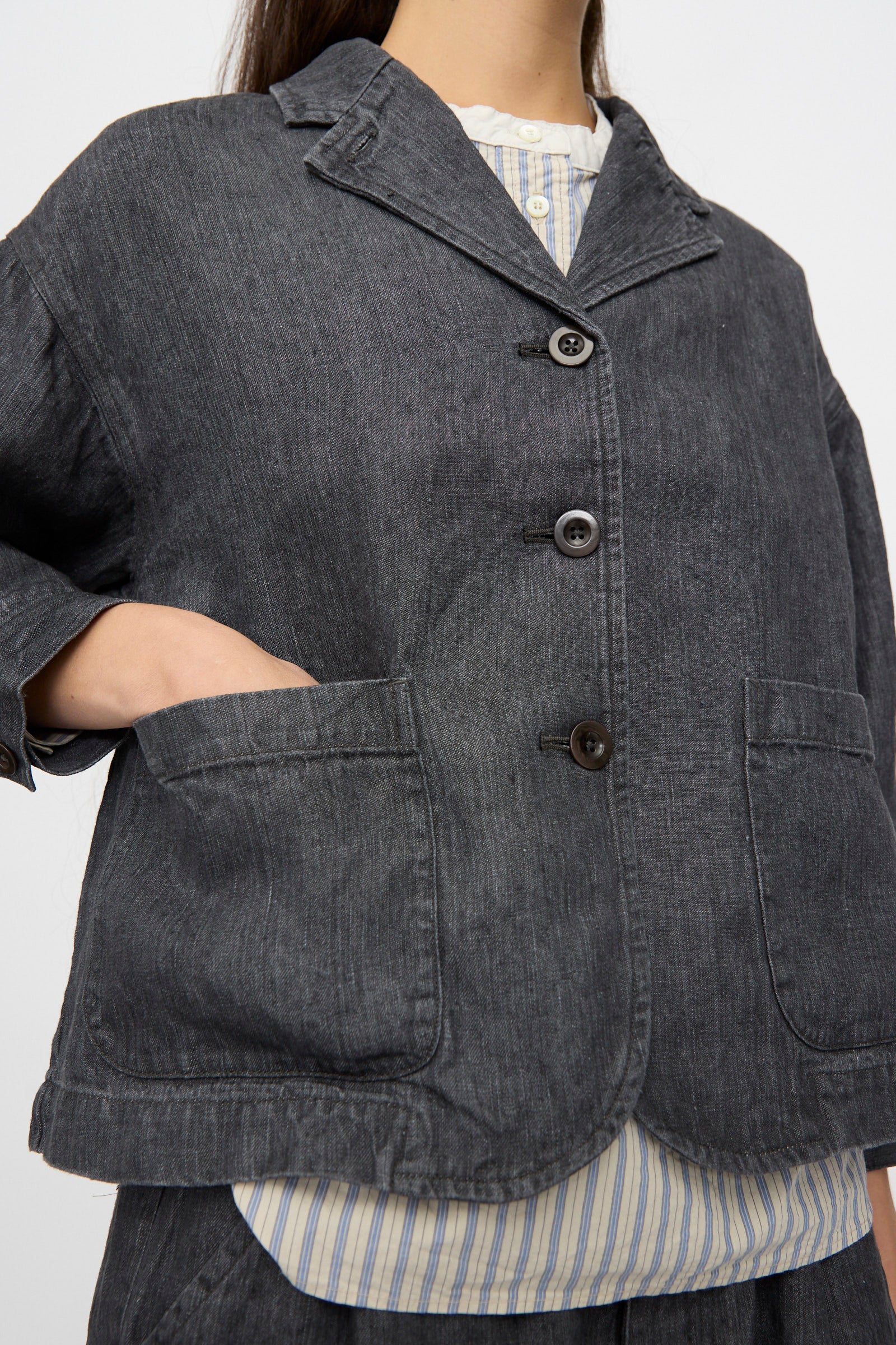 A person wears the nest Robe Linen Denim Cropped Jacket in Black, featuring a boxy fit and large front pockets, layered over a striped shirt. Only the torso and part of one arm are visible.