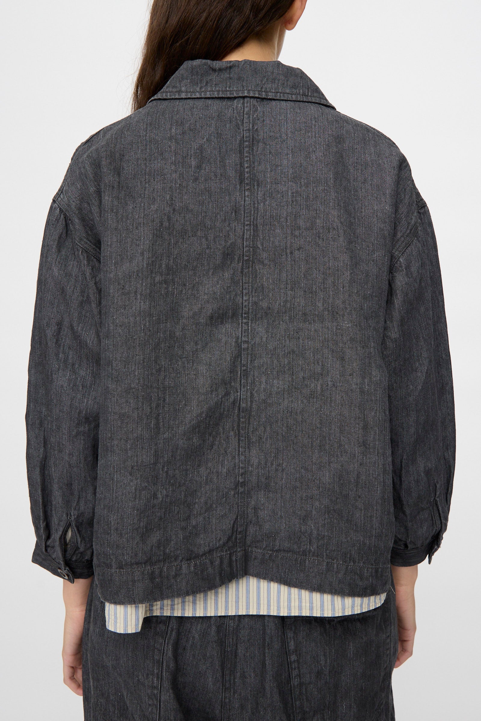 Person wearing the nest Robe Linen Denim Cropped Jacket in Black, featuring a boxy fit and long sleeves over a striped shirt. Shown from the back.