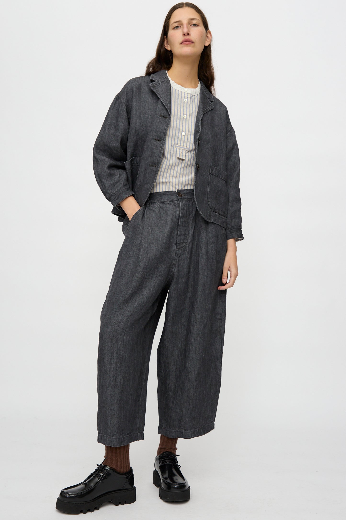 A person stands against a plain background wearing nest Robe's Linen Denim Wide Pants in Black, paired with a loose dark gray jacket, light striped shirt, brown socks, and black platform shoes.