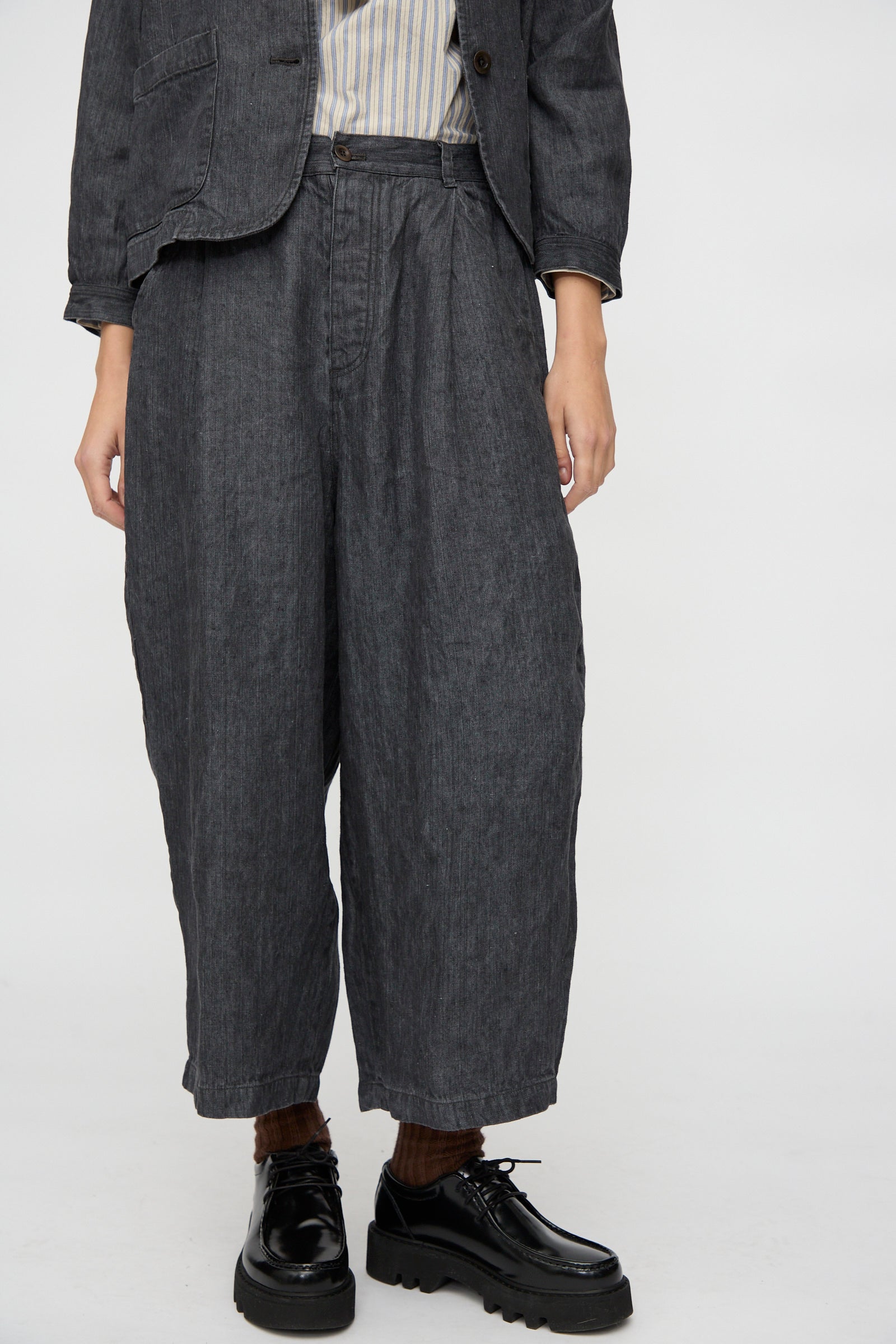 A person wears nest Robe's Linen Denim Wide Pants in Black—crafted from black linen denim and made in Japan—with a matching jacket, striped shirt, dark socks, and black platform shoes, standing against a plain background.