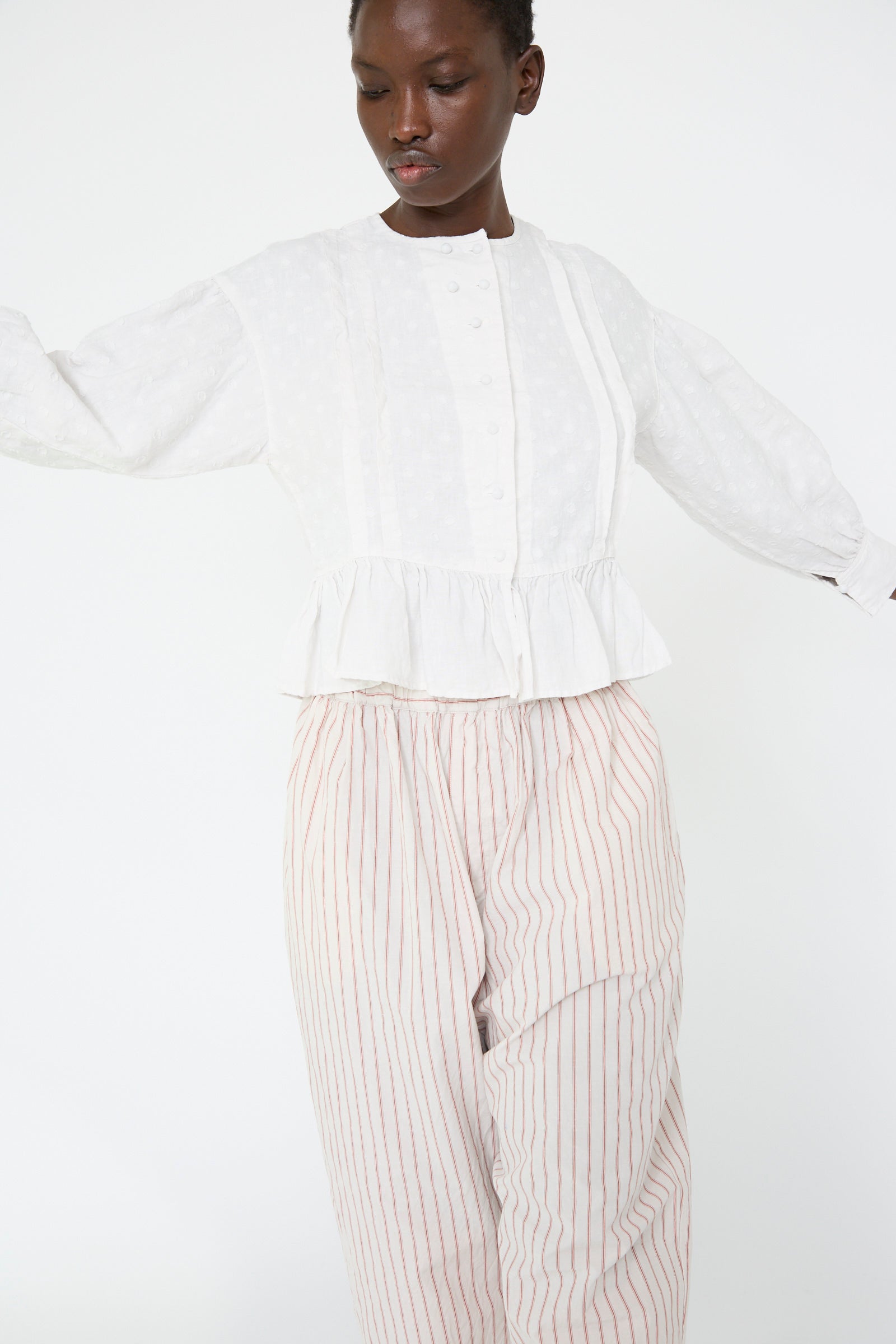 A woman wears the Linen Dot Jacquard Blouse in Off White by nest Robe and striped pants with an adjustable waist and stands against a plain white background.