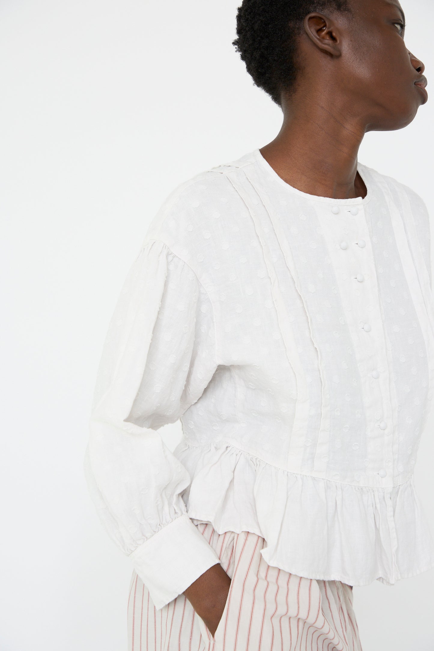 A woman wearing the Linen Dot Jacquard Blouse in Off White by nest Robe with striped pants looks away against a plain background.