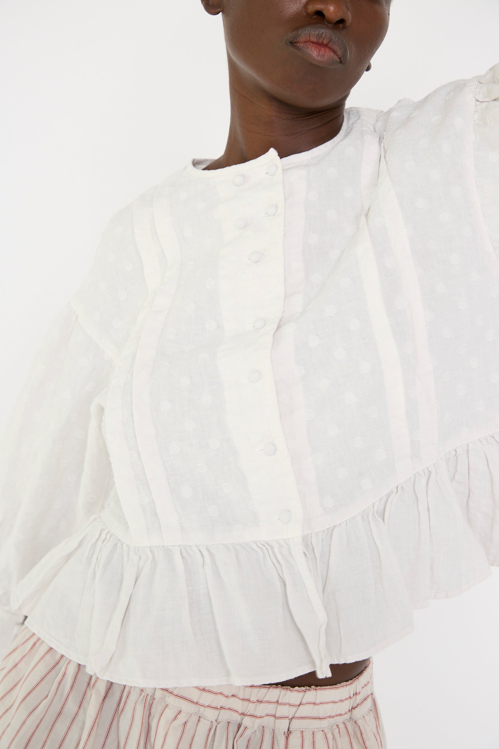 A close up of a woman standing against a plain background, wearing the Linen Dot Jacquard Blouse in Off White by nest Robe, paired with striped bottoms featuring an adjustable waist.