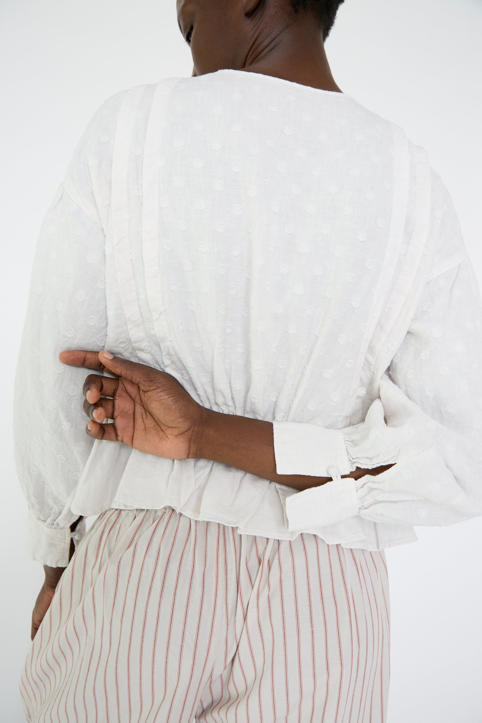 A woman wearing the Linen Dot Jacquard Blouse in Off White by nest Robe and striped pants with an adjustable waist, stands against a plain background, one hand positioned behind their back.