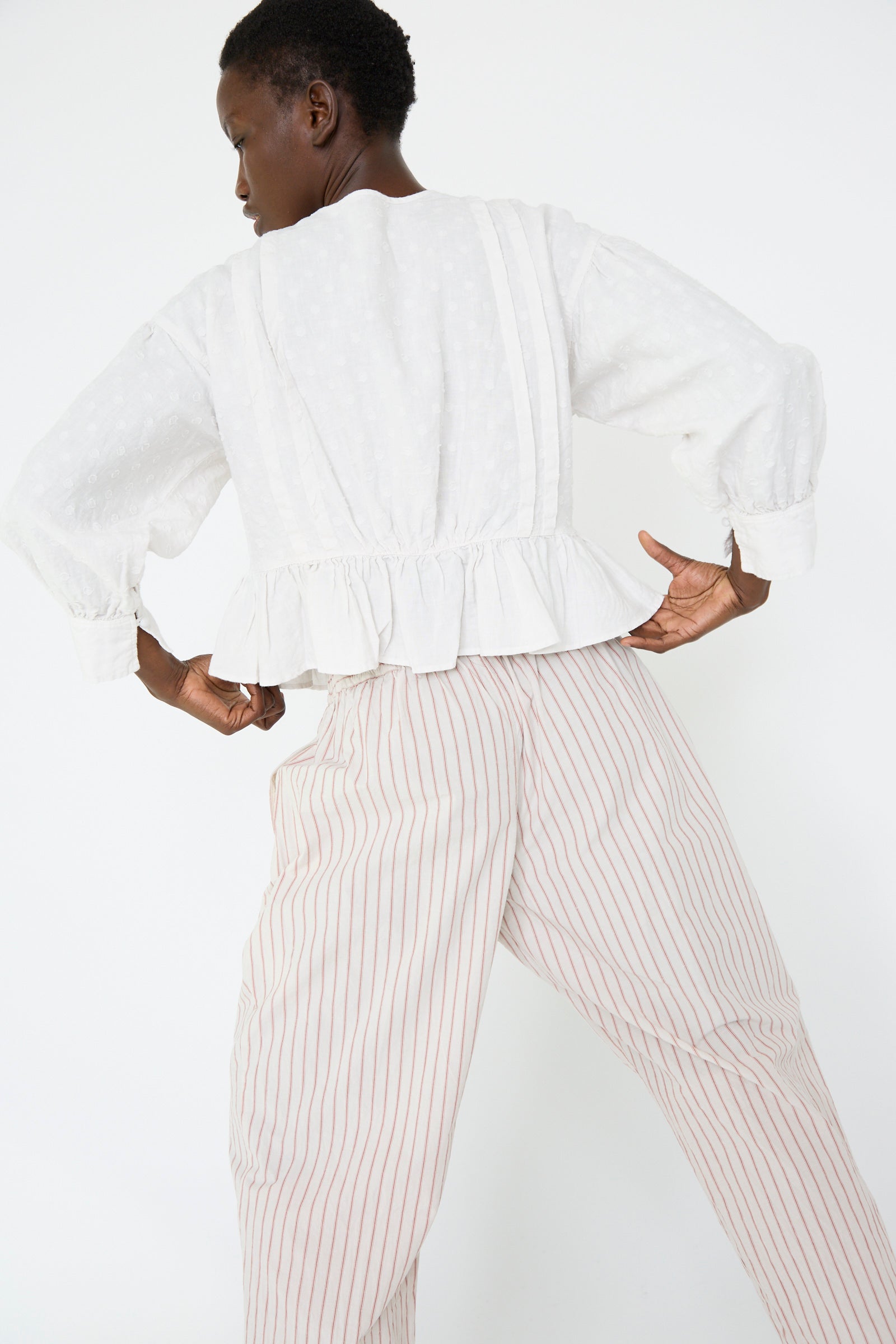 A woman wearing the Linen Dot Jacquard Blouse in Off White by nest Robe and striped pants with an adjustable waist, stands against a plain background with hands on hips.