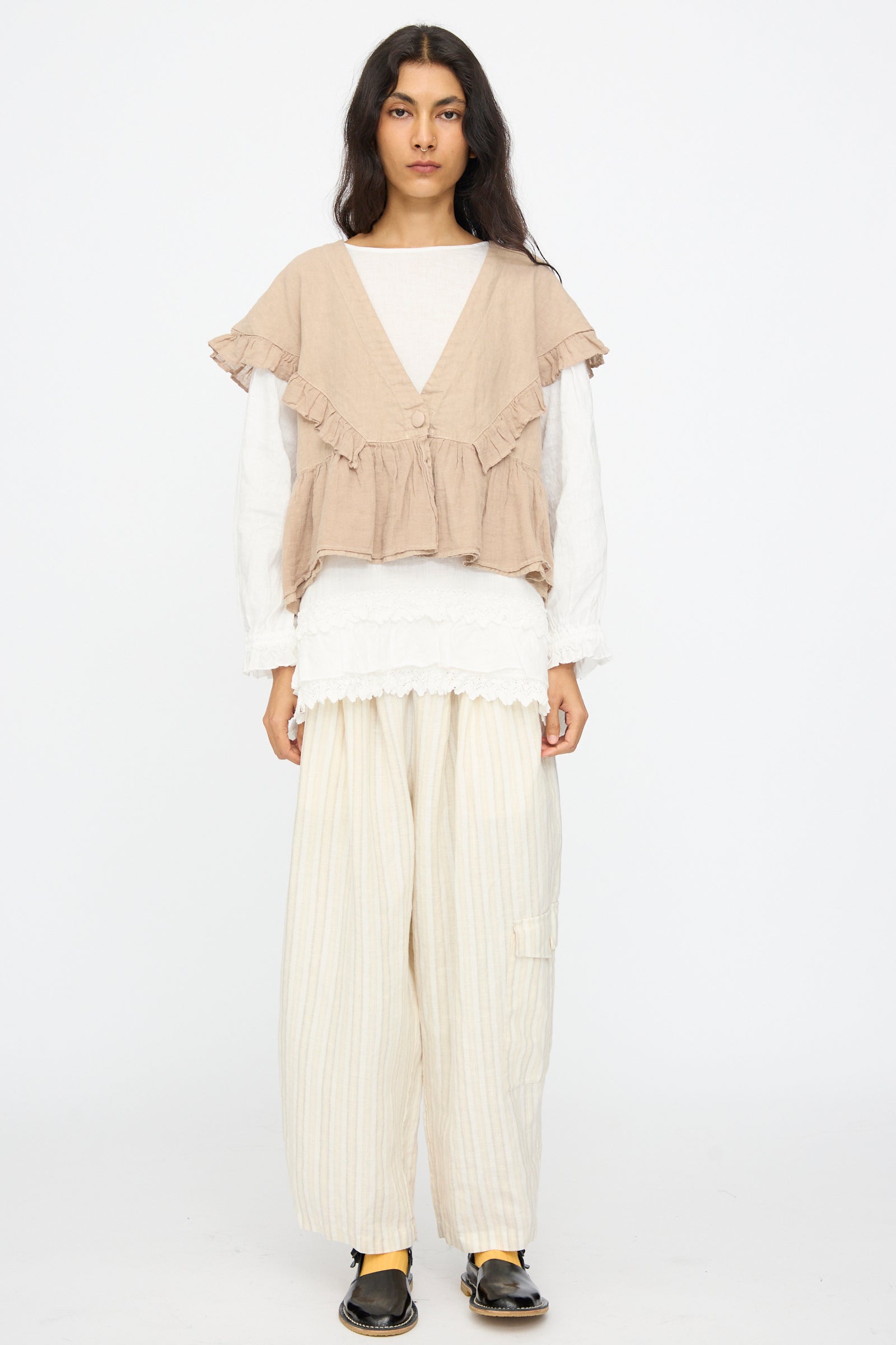 A person wearing nest Robe's Linen Easy Pants in Natural Stripe, paired with a beige ruffled vest, white long-sleeve top, and black shoes, stands against a plain white background.