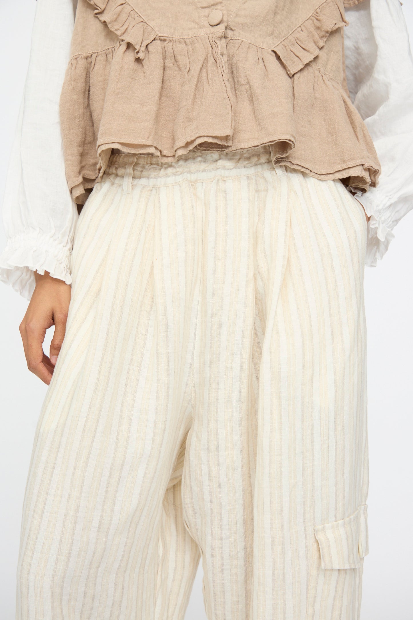 A person wears nest Robe’s Linen Easy Pants in Natural Stripe, paired with a beige ruffled top over a white long-sleeved shirt; only their torso and upper legs are shown.