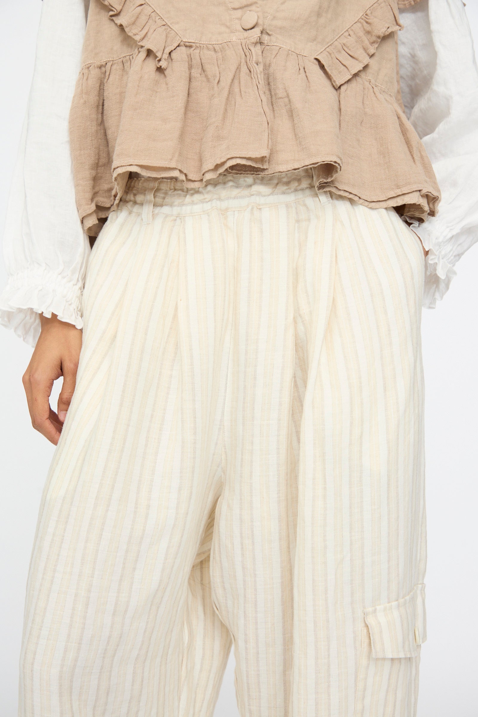 A person wears nest Robe’s Linen Easy Pants in Natural Stripe, paired with a beige ruffled top over a white long-sleeved shirt; only their torso and upper legs are shown.