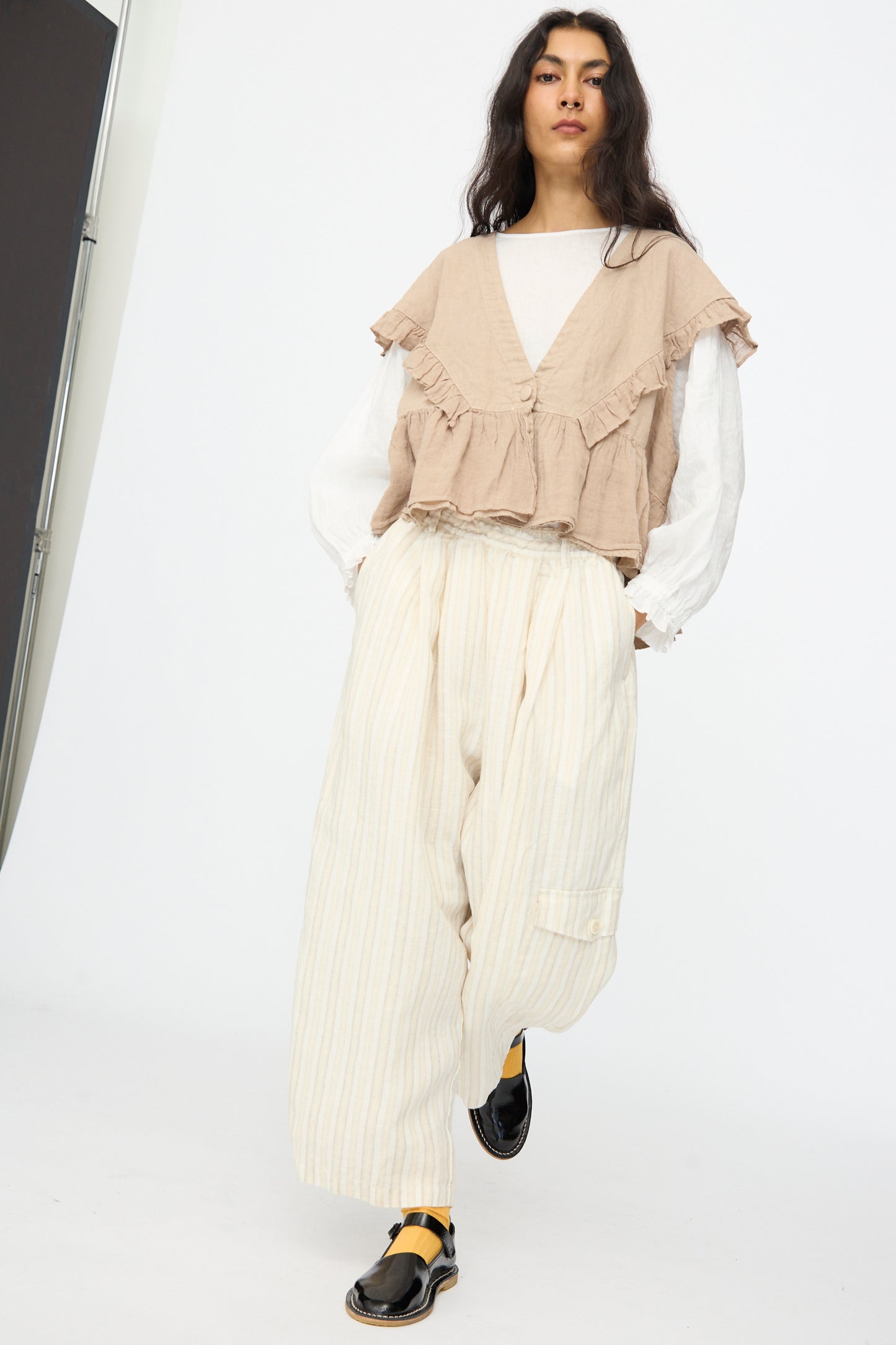A person wears nest Robe Linen Easy Pants in Natural Stripe with a beige ruffled vest, a white long-sleeve top, and black and yellow shoes, standing against a plain white background.