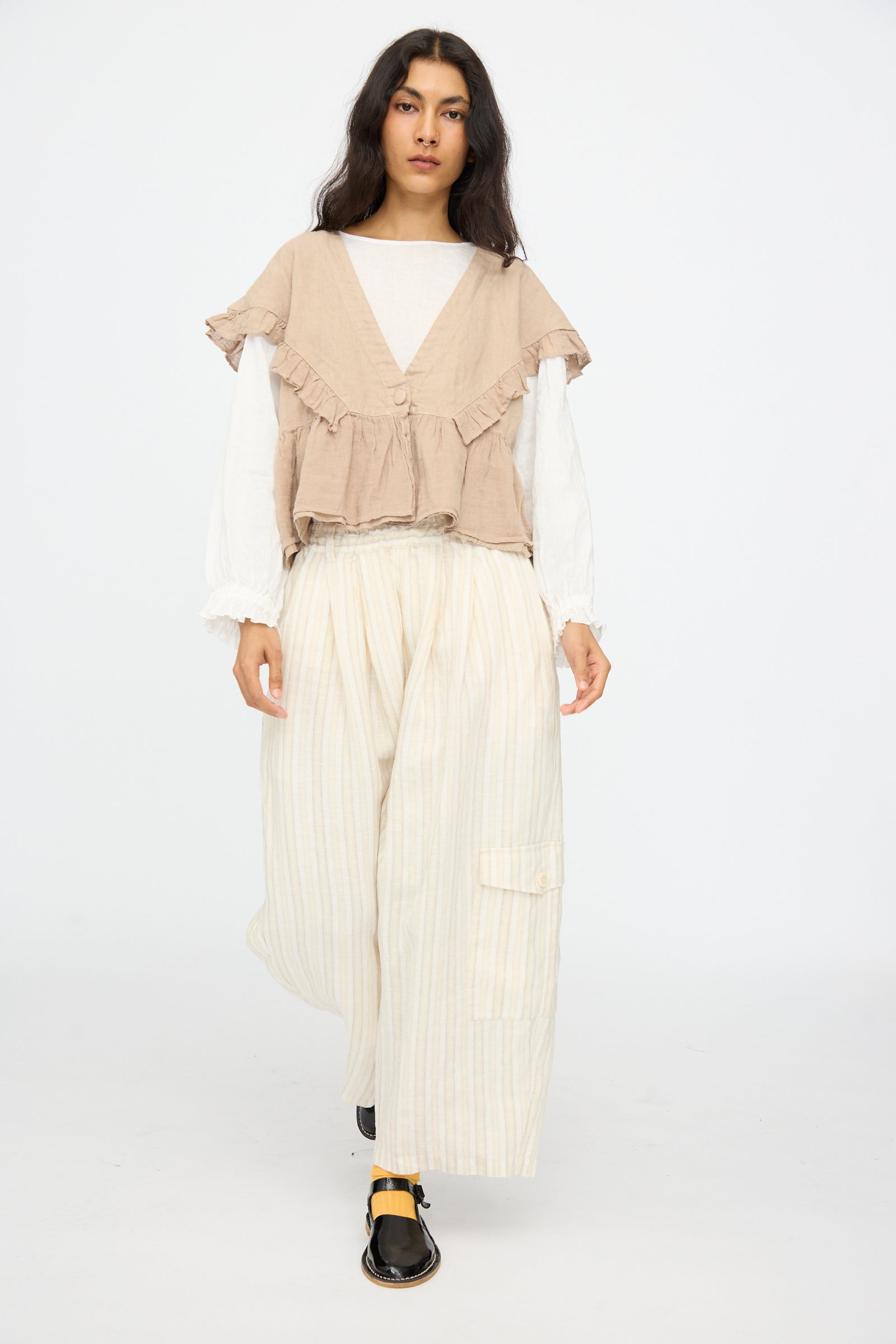 A person with long dark hair wears nest Robe's Linen Easy Pants in Natural Stripe, paired with a beige ruffled vest over a white long-sleeve top, walking against a plain white background.