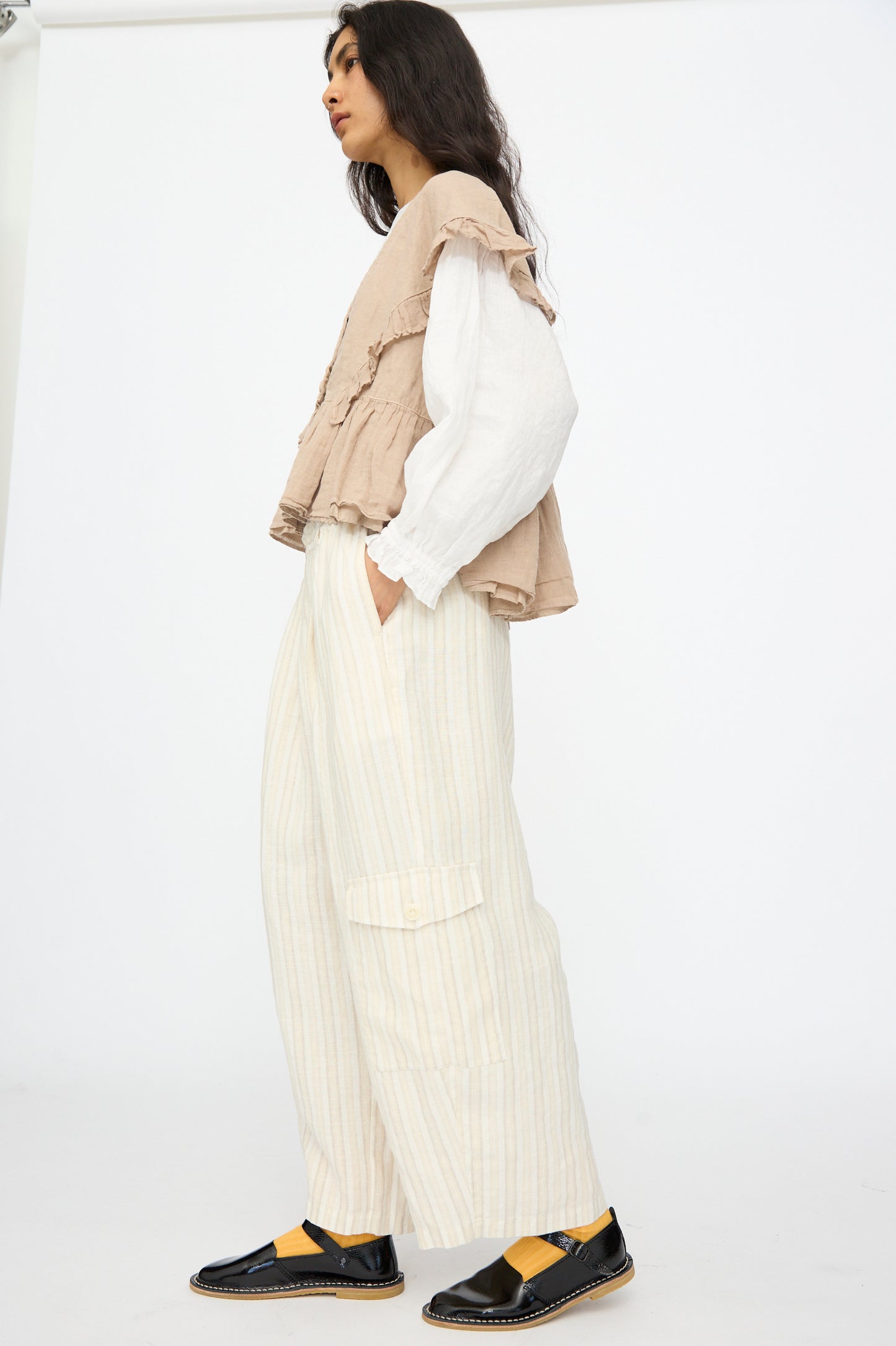 A person stands sideways against a plain background wearing nest Robe’s Linen Easy Pants in Natural Stripe, featuring an elastic waistband, paired with a beige ruffled vest, white long-sleeve shirt, and black shoes with yellow socks.
