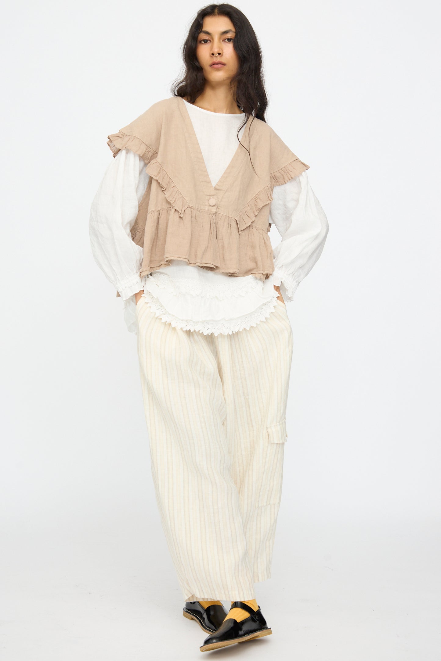 A woman stands against a white background wearing nest Robe's Linen Easy Pants in Natural Stripe, paired with a beige ruffled vest, white long-sleeve top, black sandals, and yellow socks.