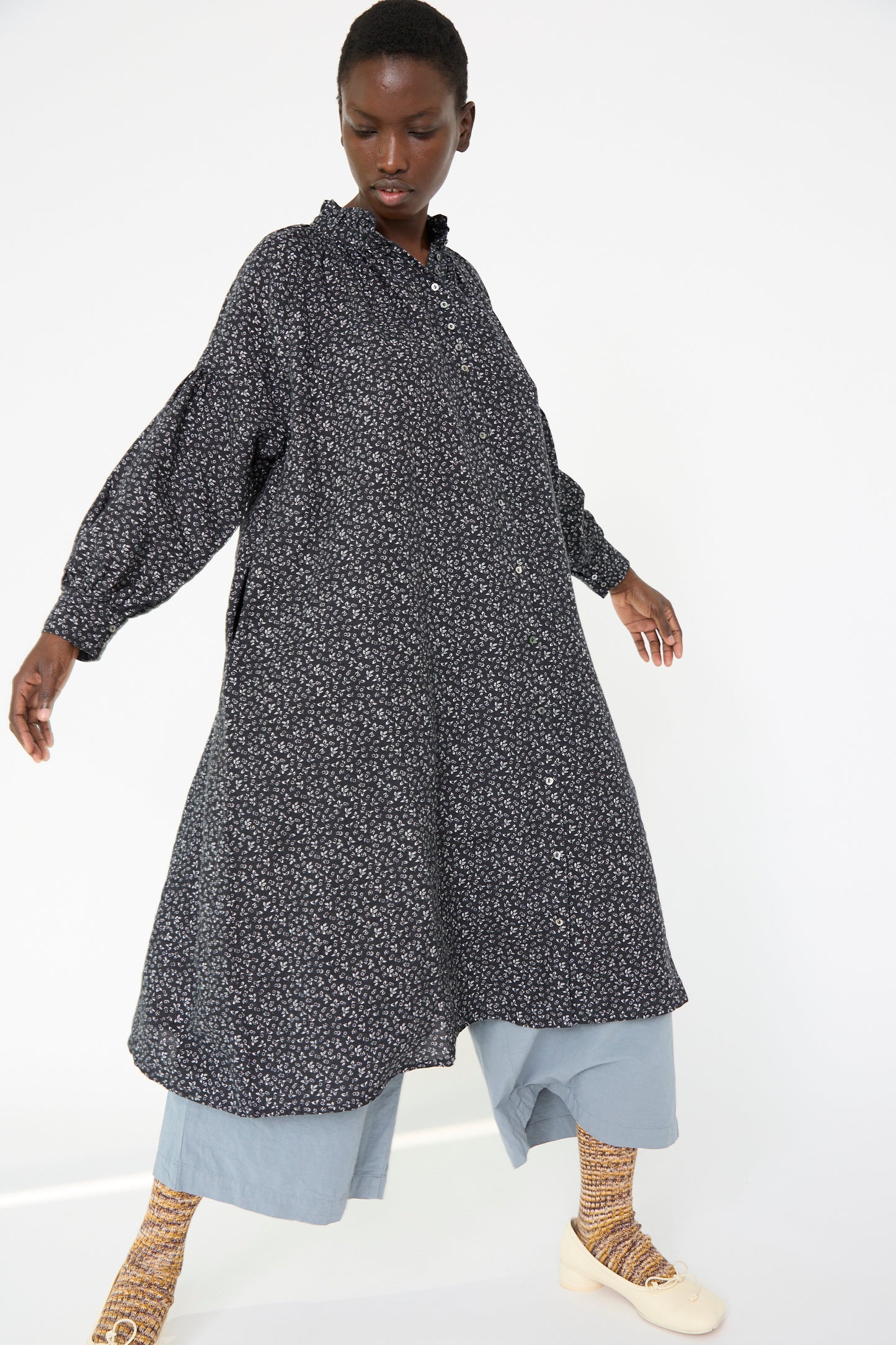 A person wearing the Linen Floral Print Dress in Black by nest Robe over light blue pants and patterned socks with beige shoes, posing against a plain white background.