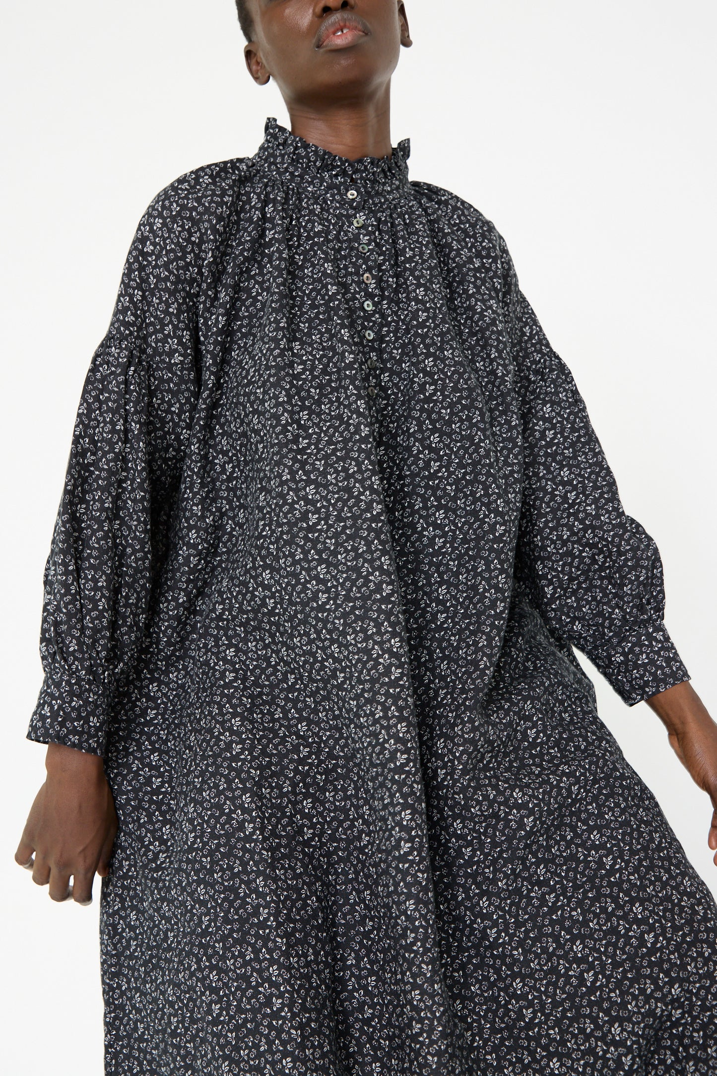 A person wearing the Linen Floral Print Dress in Black by nest Robe, featuring a high collar and buttoned front, stands against a plain background.