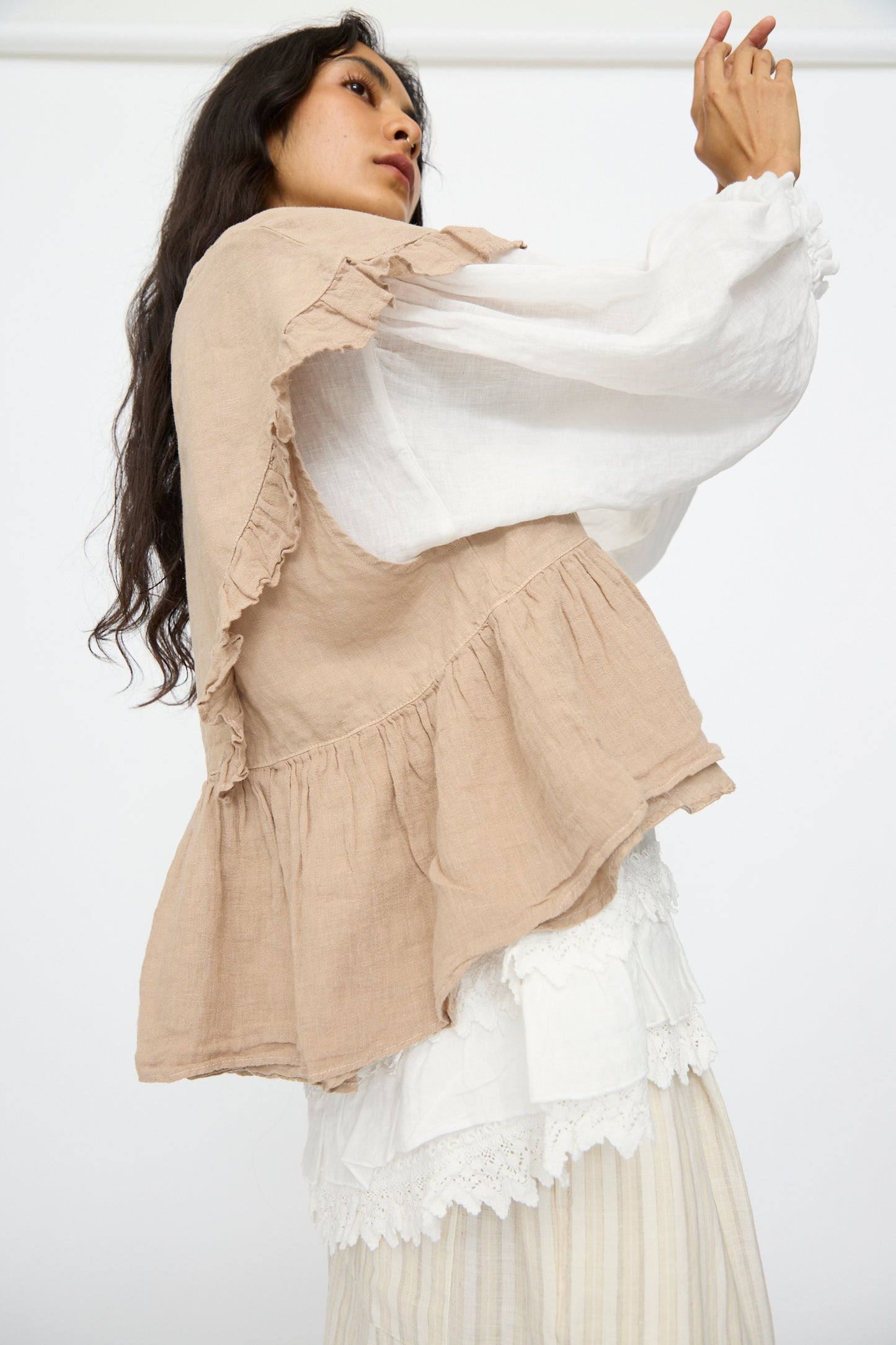 A model wears the nest Robe Linen Frill Vest in Beige over a white long-sleeve blouse and pleated skirt, standing against a plain white background.