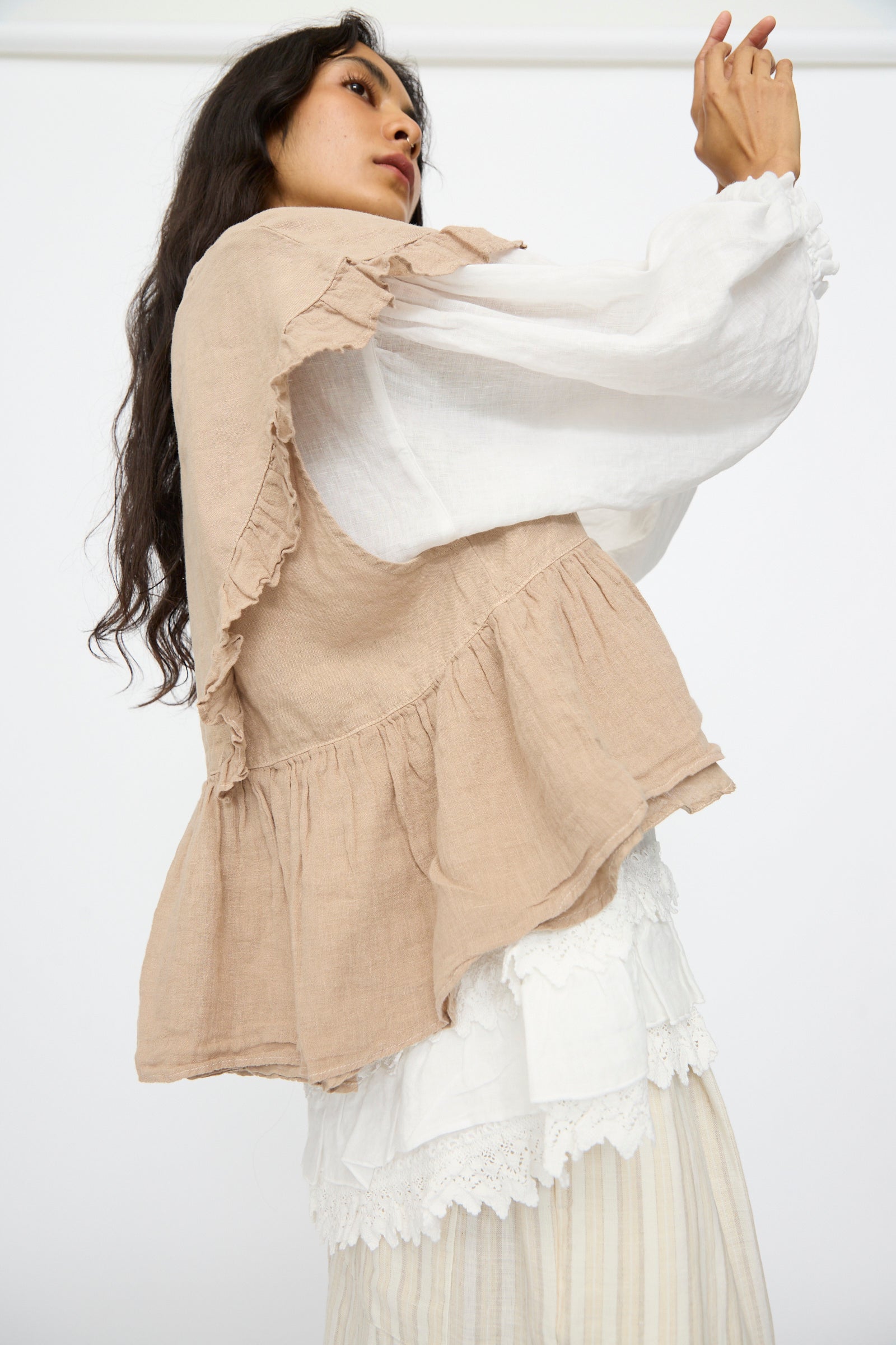 A model wears the nest Robe Linen Frill Vest in Beige over a white long-sleeve blouse and pleated skirt, standing against a plain white background.