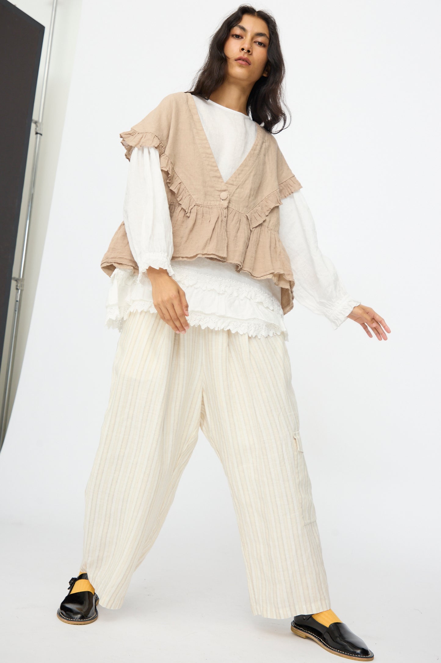 A person stands indoors against a plain background wearing nest Robe's Linen Frill Vest in Beige, loose cream-colored striped pants, a white layered top, and black slip-on shoes.