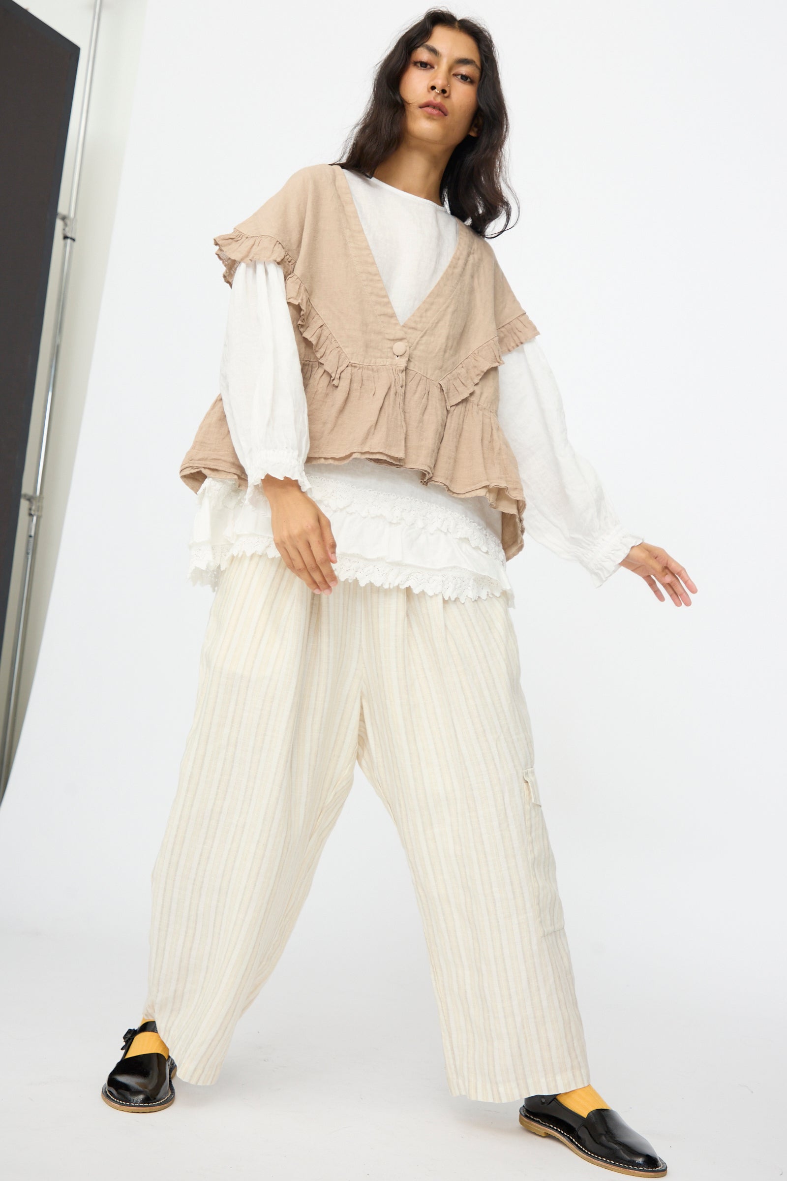 A person stands indoors against a plain background wearing nest Robe's Linen Frill Vest in Beige, loose cream-colored striped pants, a white layered top, and black slip-on shoes.