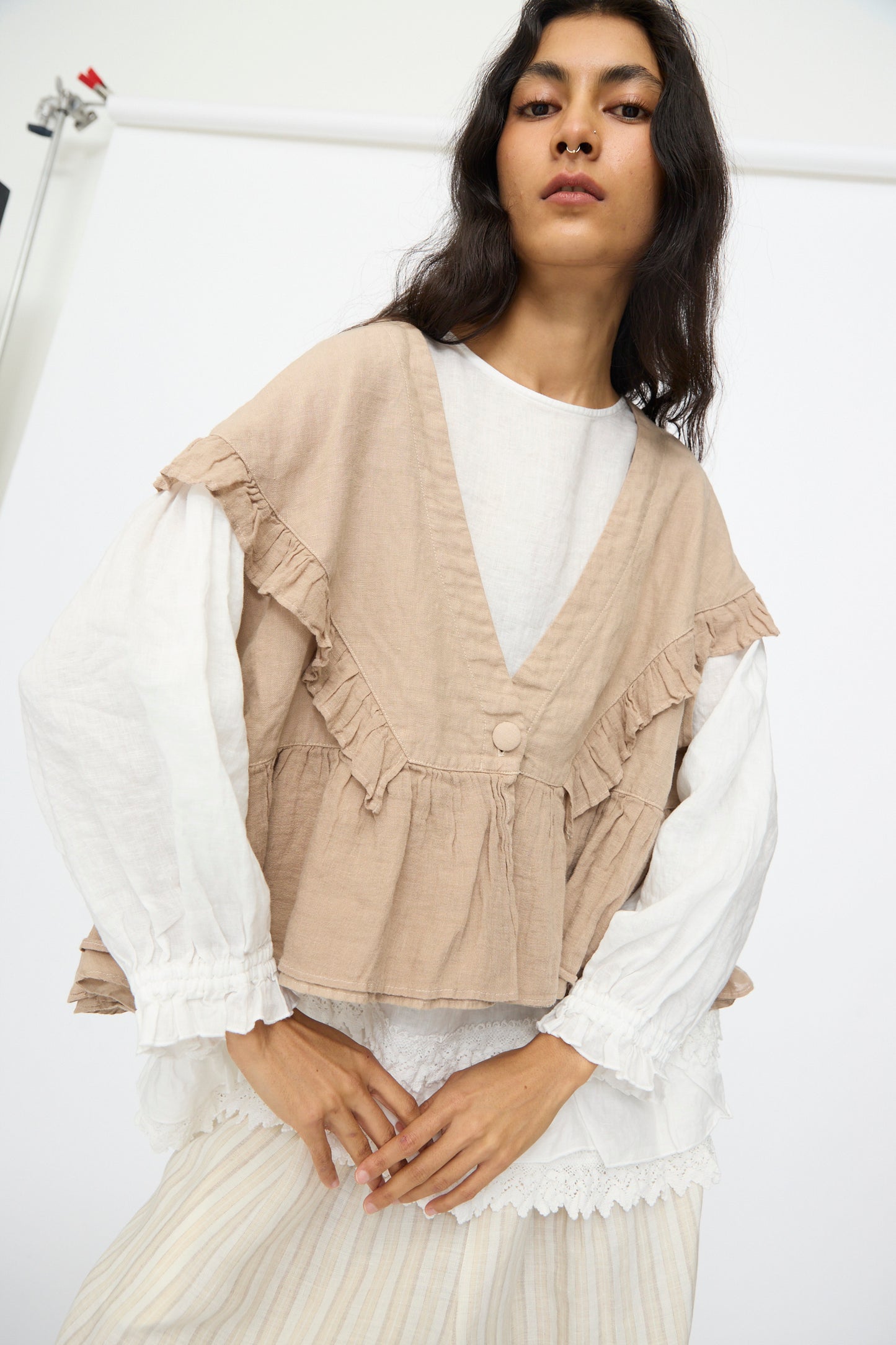 A woman wears the nest Robe Linen Frill Vest in Beige over a white long-sleeve blouse and a light skirt, standing against a plain white background.