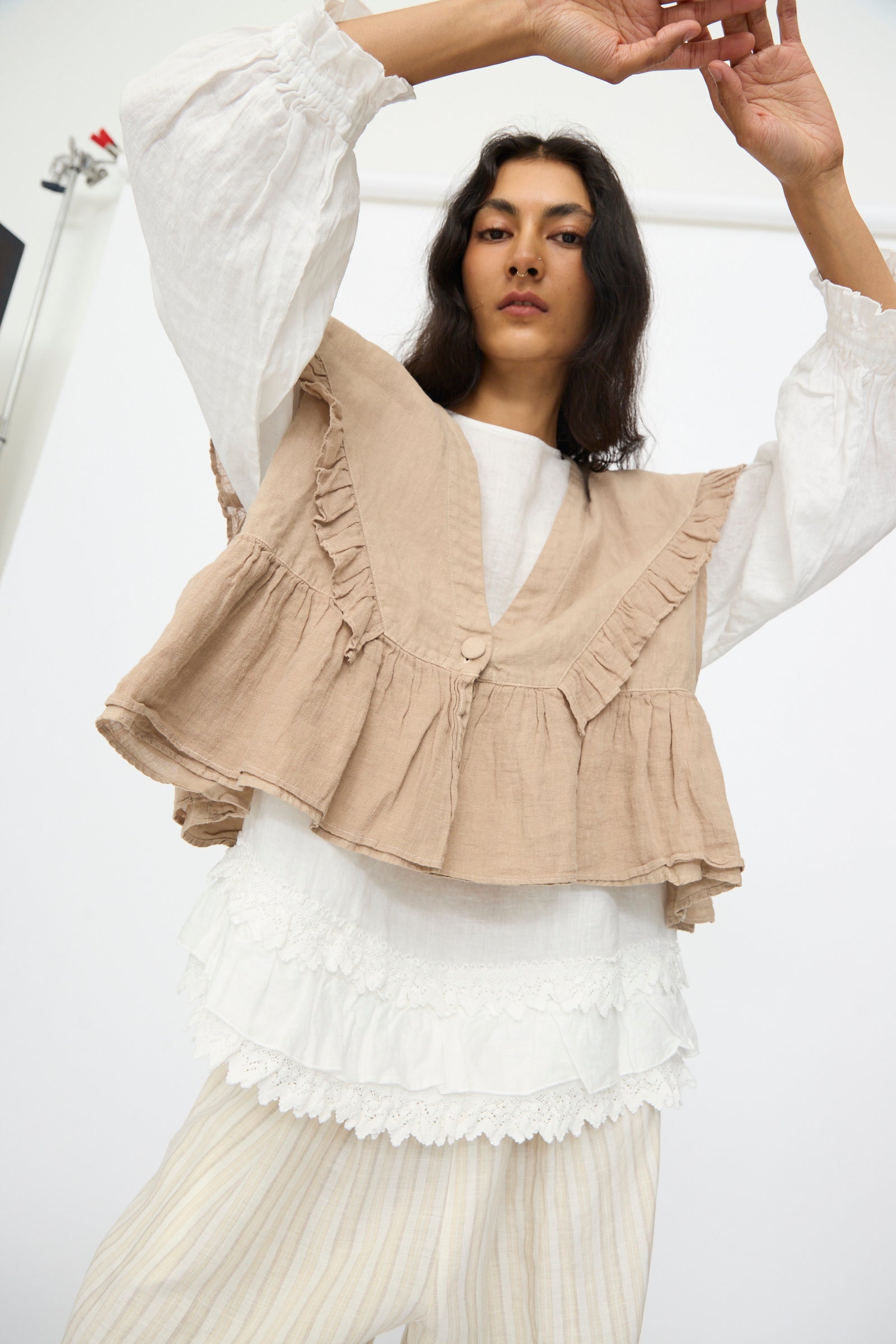 A woman wears the nest Robe Linen Frill Vest in Beige over a white blouse and striped pants, standing with arms raised against a plain background.