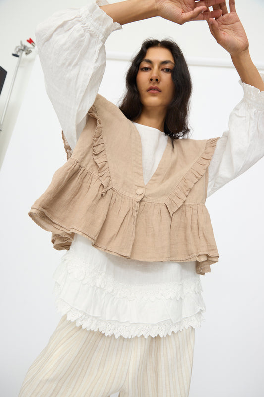 A woman wears the nest Robe Linen Frill Vest in Beige over a white blouse and striped pants, standing with arms raised against a plain background.