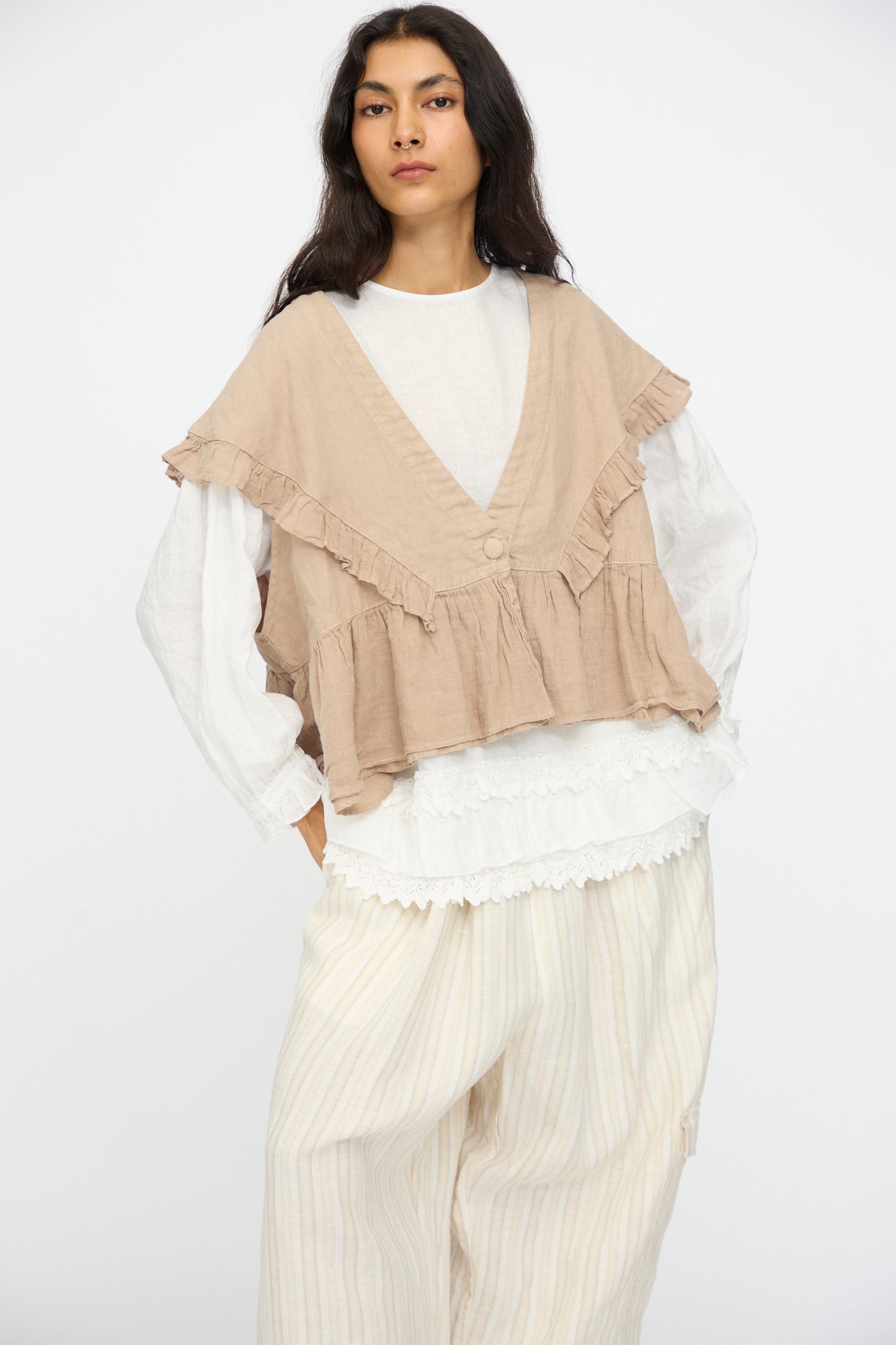 A person wears the nest Robe Linen Frill Vest in Beige over a white long-sleeve top and loose light-striped pants, standing against a plain white background.