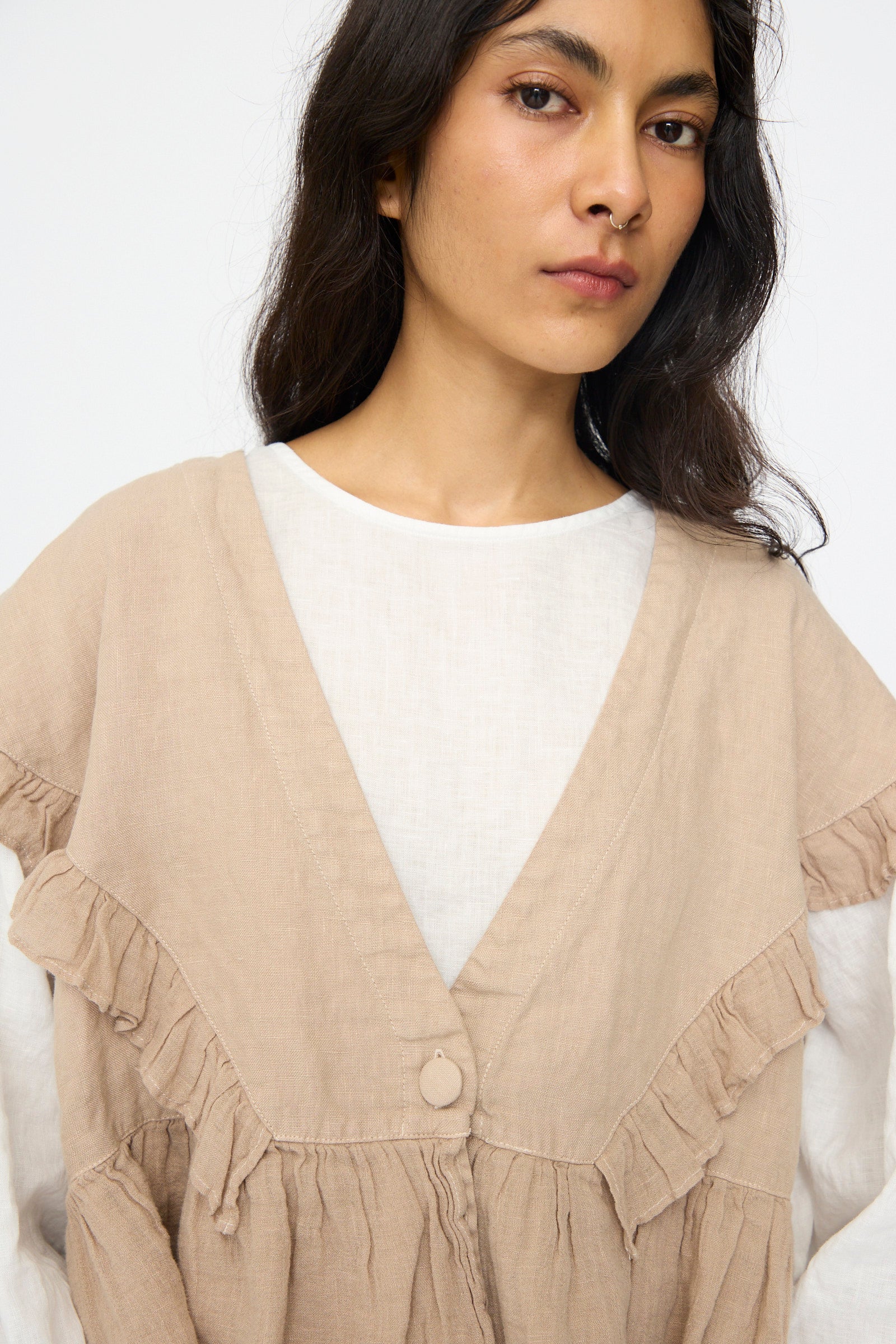 A woman with long dark hair models the Linen Frill Vest in Beige from nest Robe over a white top, standing against a plain light background.