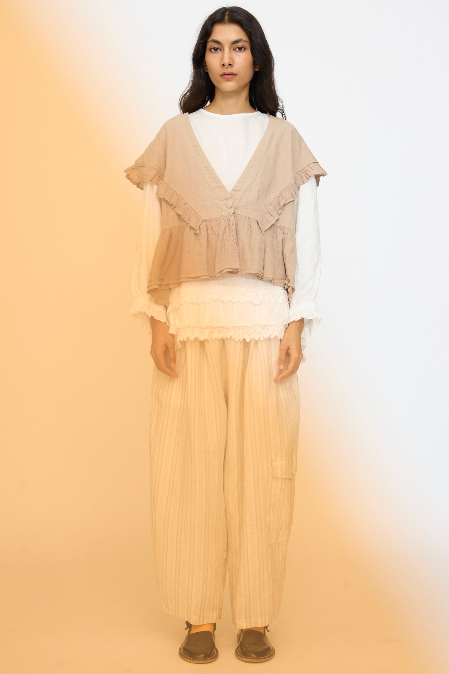 A person faces forward against a soft orange gradient background, wearing the nest Robe Linen Frill Vest in Beige over a white long-sleeve top, with loose beige pants and brown shoes.