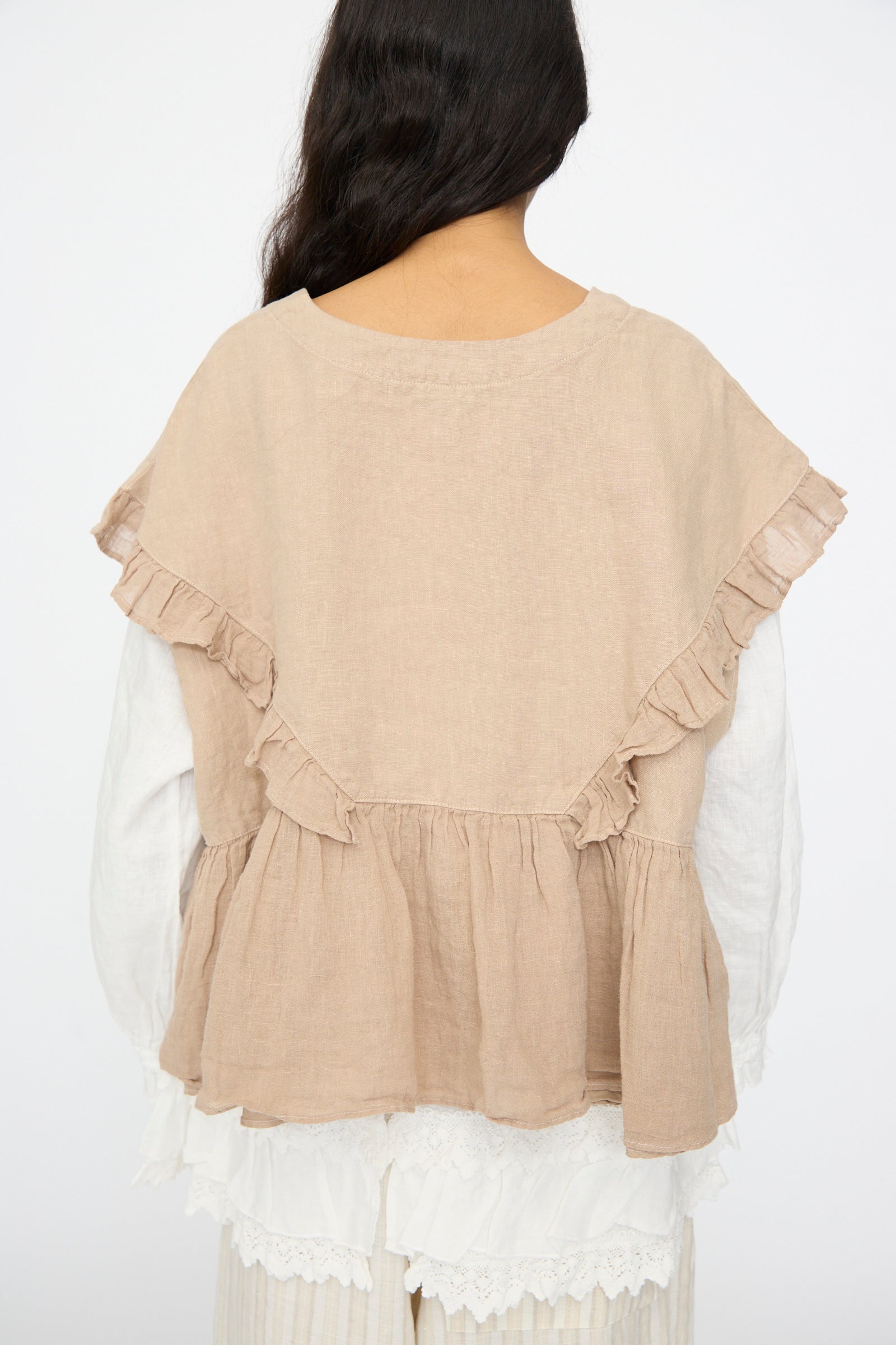 A person with long dark hair is photographed from the back against a plain background, wearing the nest Robe Linen Frill Vest in Beige layered over a white blouse.