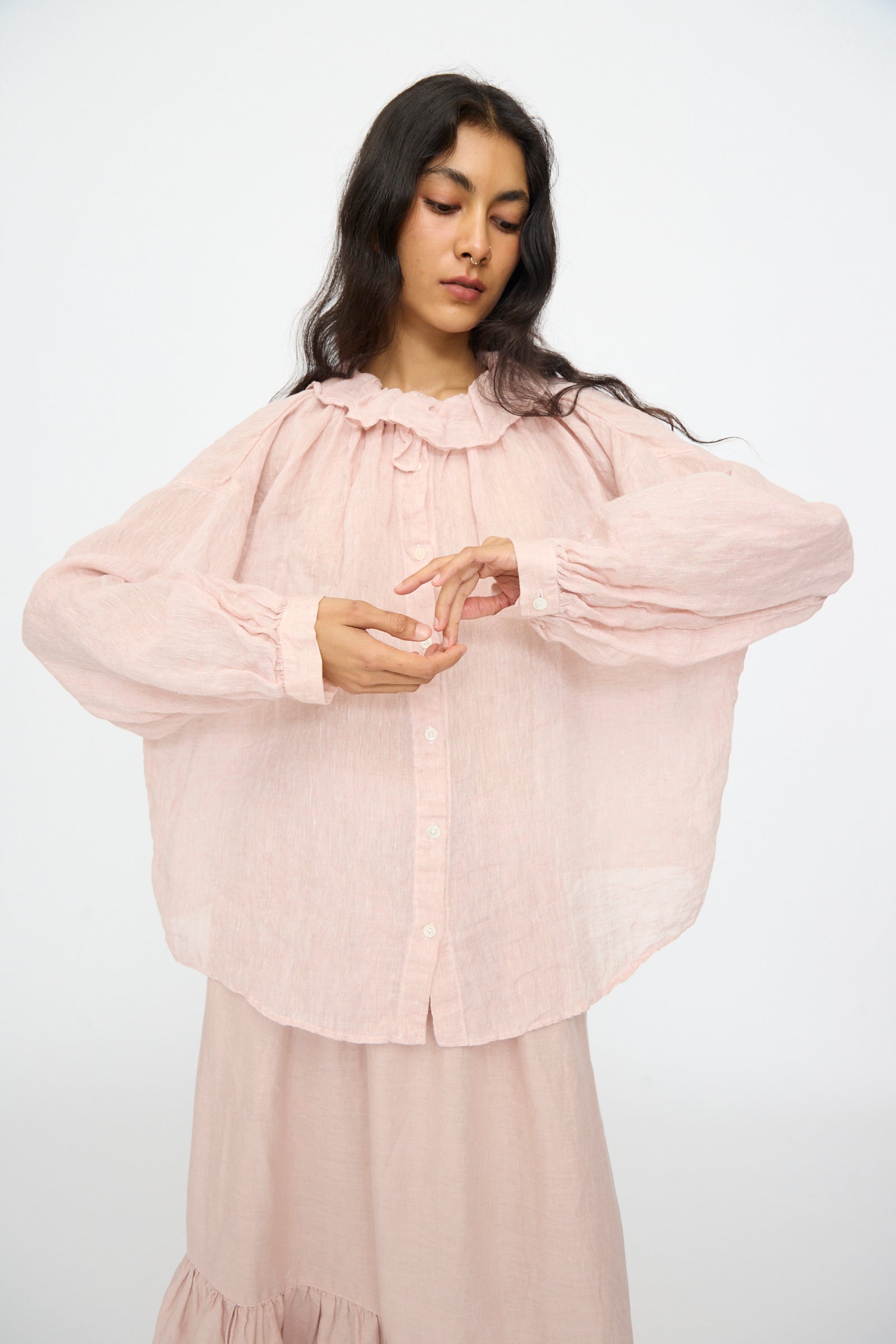 A woman in the nest Robe Linen Gauze Frill Blouse in Light Peach stands against a plain background, looking down with her hands gently touching.