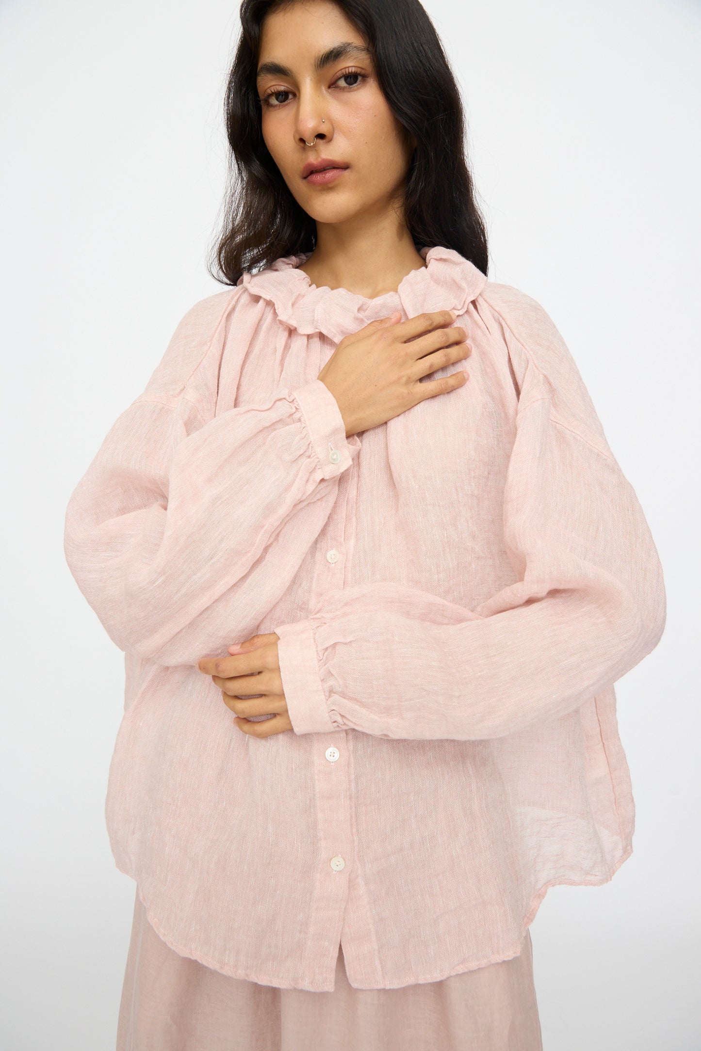 A person models the nest Robe Linen Gauze Frill Blouse in Light Peach, a loose-fit linen top with long sleeves and a ruffle collar, standing against a plain background with one hand on their chest and one resting on their torso.