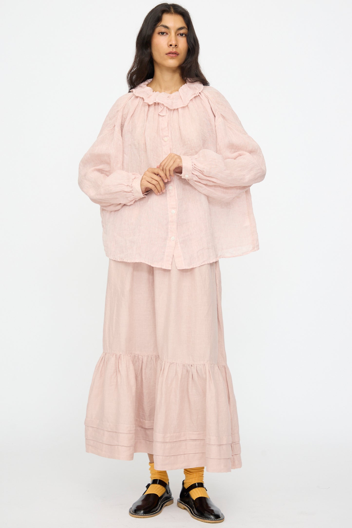 A person stands facing forward, wearing the nest Robe Linen Gauze Frill Blouse in Light Peach with a pink tiered skirt, yellow socks, and black shoes.