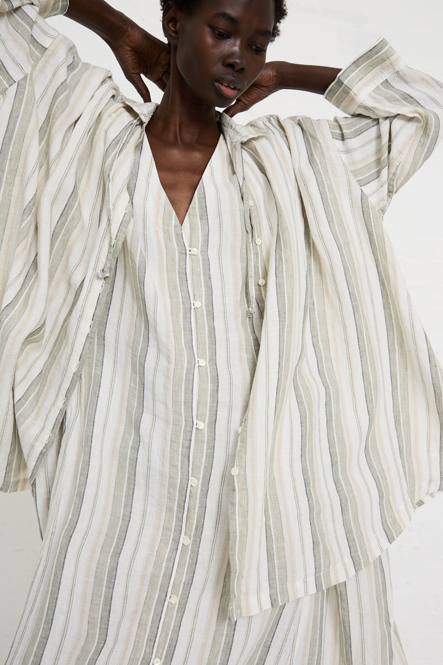 A person wearing a loose-fitting, striped **Linen Long Oversized Shirt in Stripe** by **nest Robe** with a matching striped dress underneath. They have their hands raised, adjusting the collar of the relaxed fit outer garment.