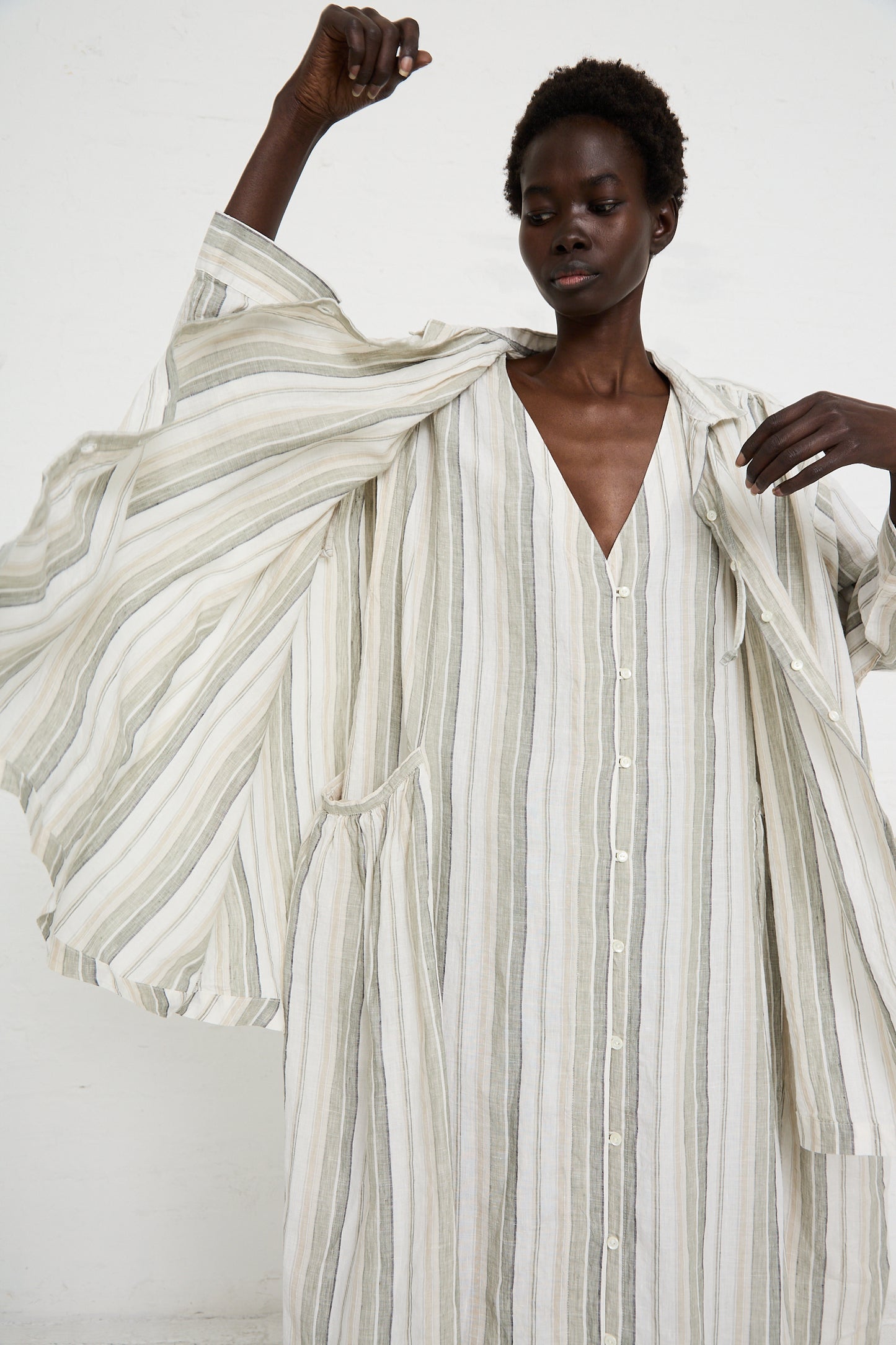 Person poses in a Linen Long Oversized Shirt in Stripe by nest Robe, lifting one arm up to reveal wide, flowing sleeves. The dress embodies a minimalist, neutral color palette with a relaxed fit perfect for any occasion.