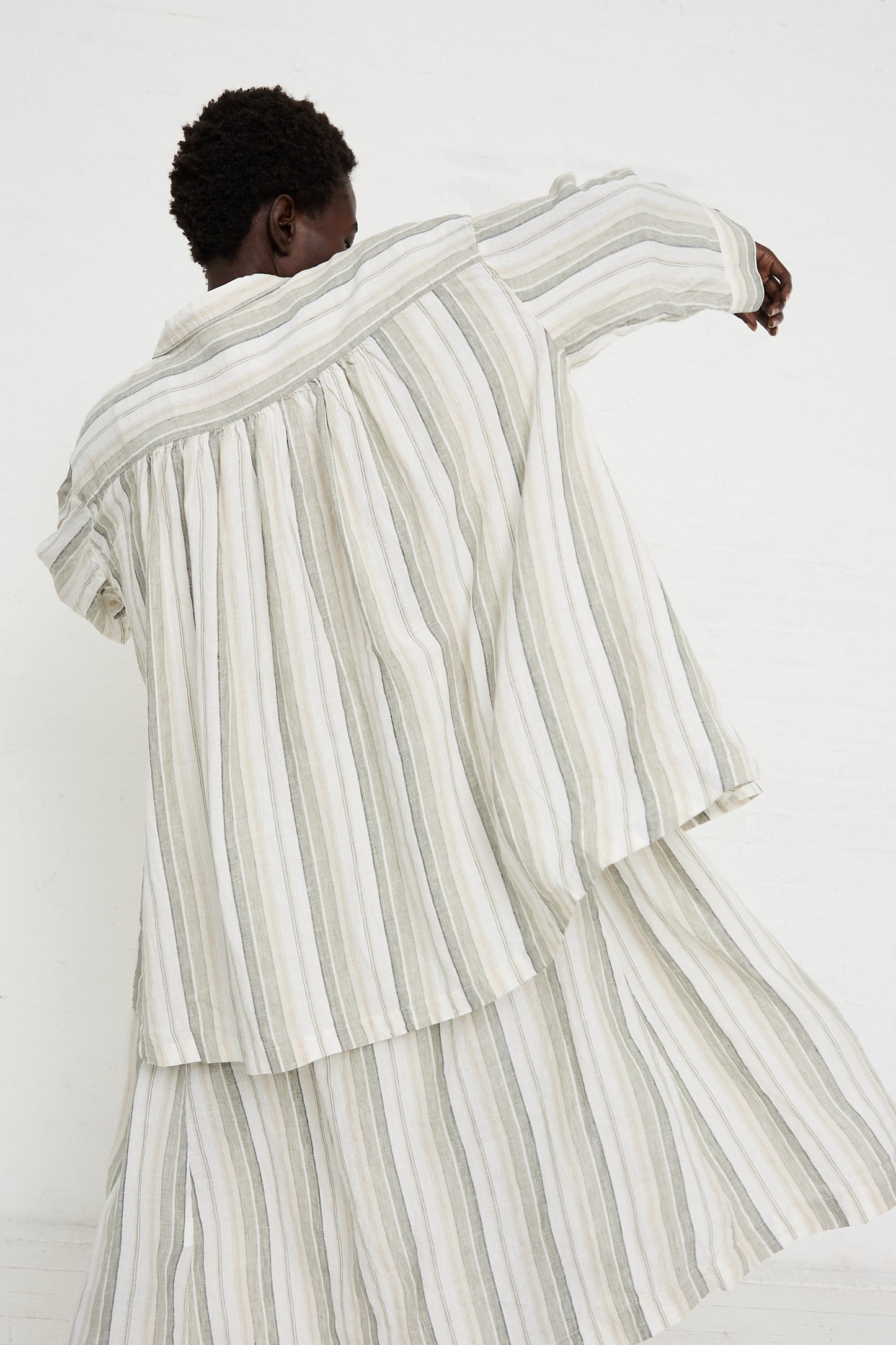 Person wearing a Linen Long Oversized Shirt in Stripe by nest Robe, with their back facing the camera, raising one arm slightly. The relaxed fit attire features flowing, vertical stripes in light and dark shades.