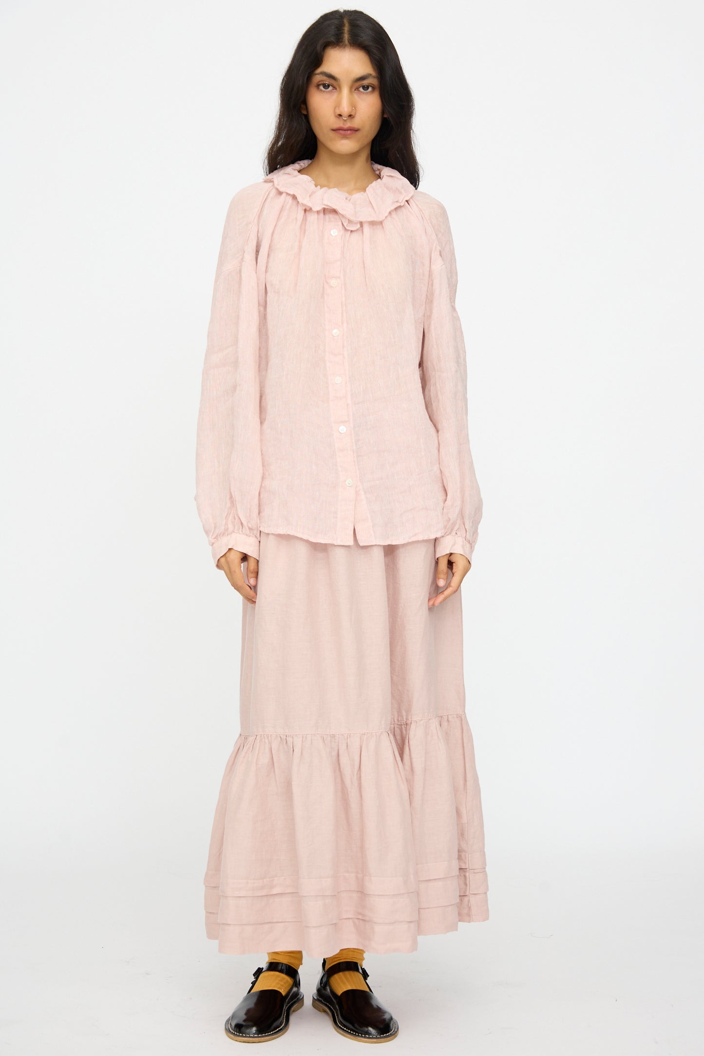 A woman with dark hair faces forward, wearing a light pink long-sleeve blouse and the nest Robe Linen Natural Dyed Pleated Gathered Skirt in Pink. She pairs the outfit with black shoes and yellow socks.