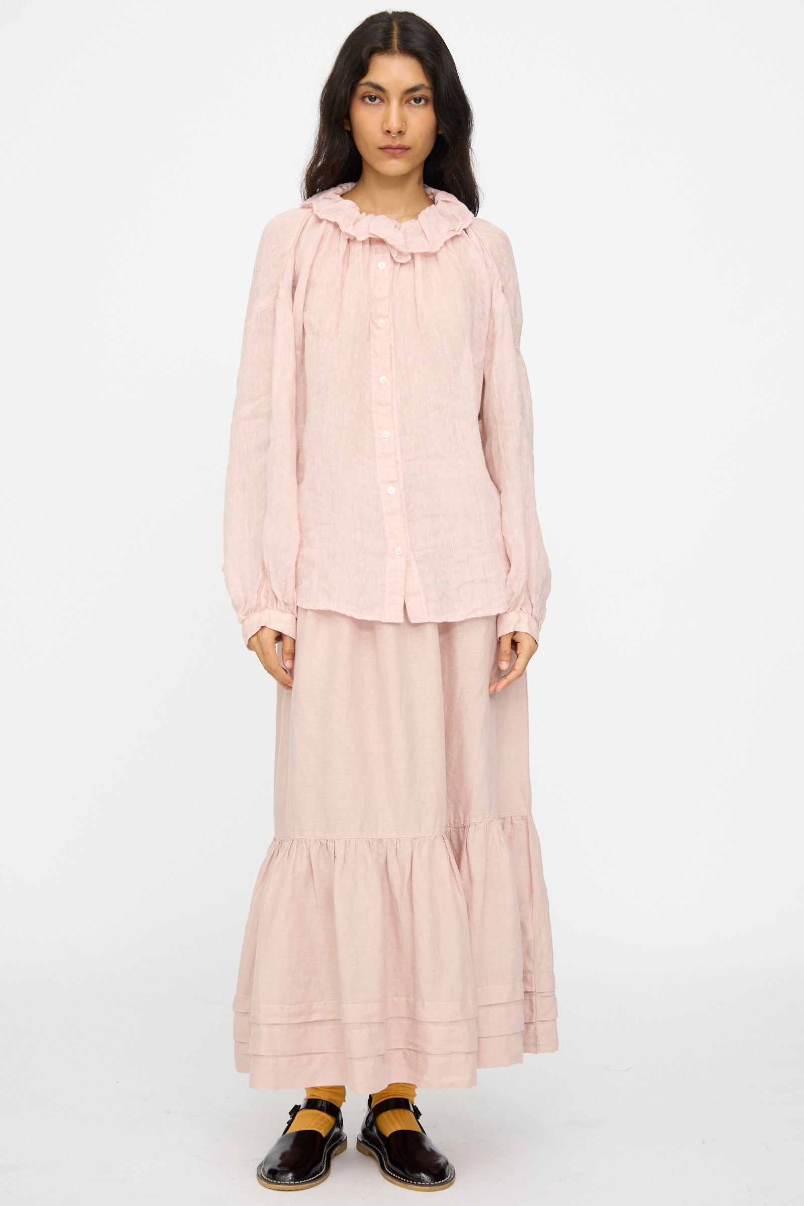 A woman with dark hair faces forward, wearing a light pink long-sleeve blouse and the nest Robe Linen Natural Dyed Pleated Gathered Skirt in Pink. She pairs the outfit with black shoes and yellow socks.