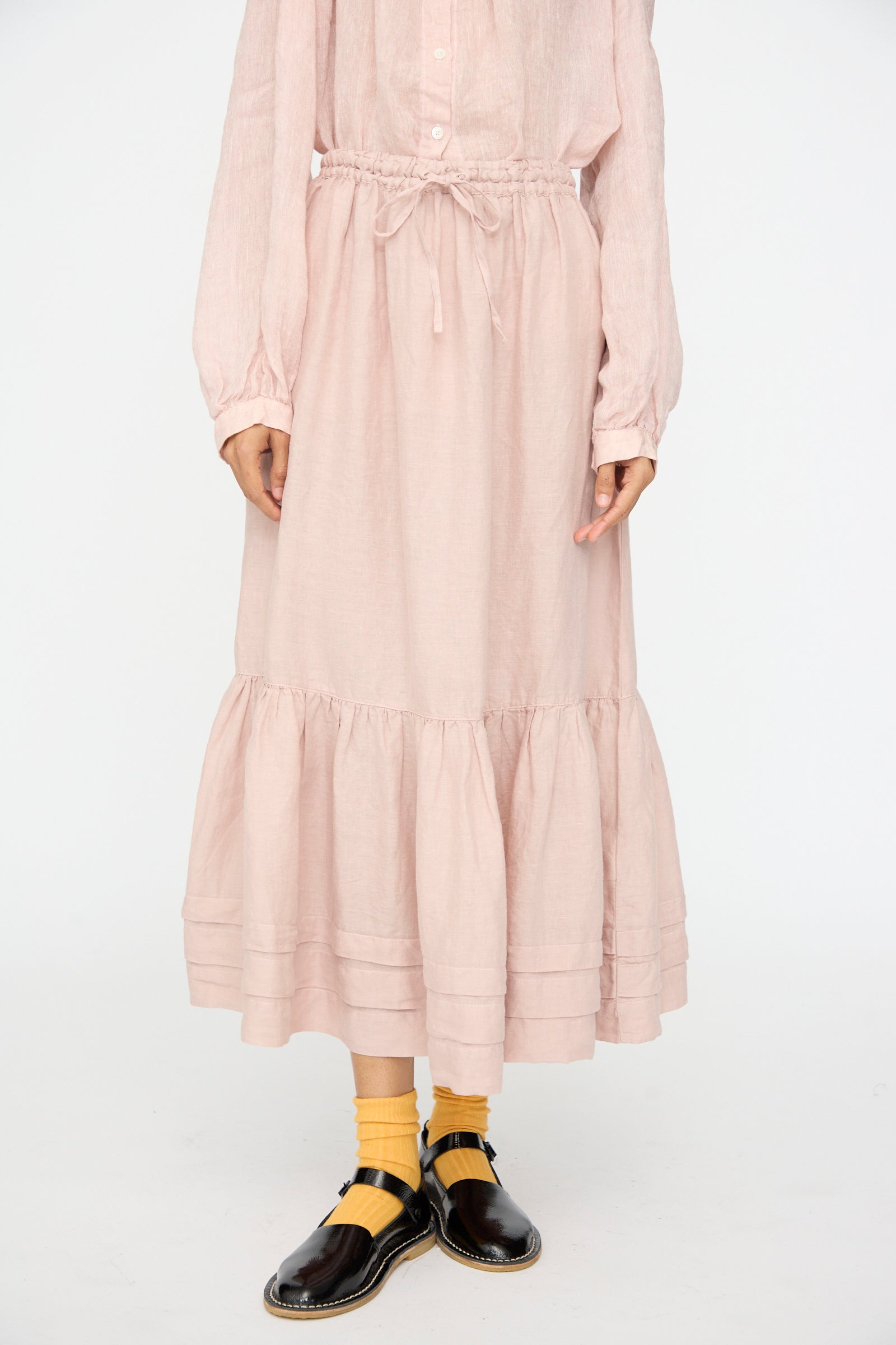 A person wearing the nest Robe Linen Natural Dyed Pleated Gathered Skirt in Pink with a matching light pink blouse, yellow socks, and black shoes stands against a plain white background.